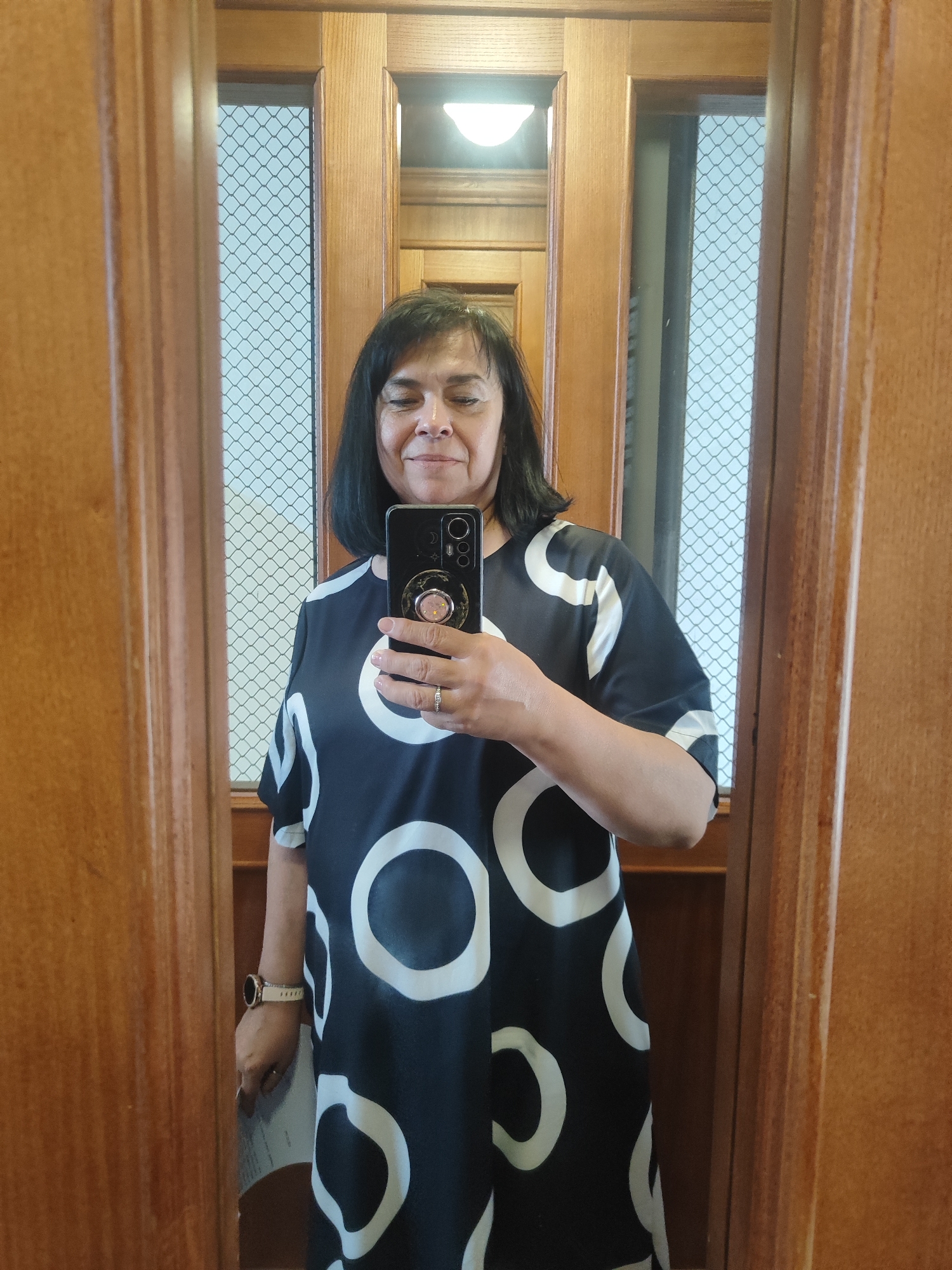 A woman stands in a doorway, taking a mirror selfie. She is wearing a black dress with a white circle pattern, a watch, and a ring. She holds a smartphone in her right hand. The environment appears to be indoors, with wooden paneling and a window with a grid pattern visible in the background.