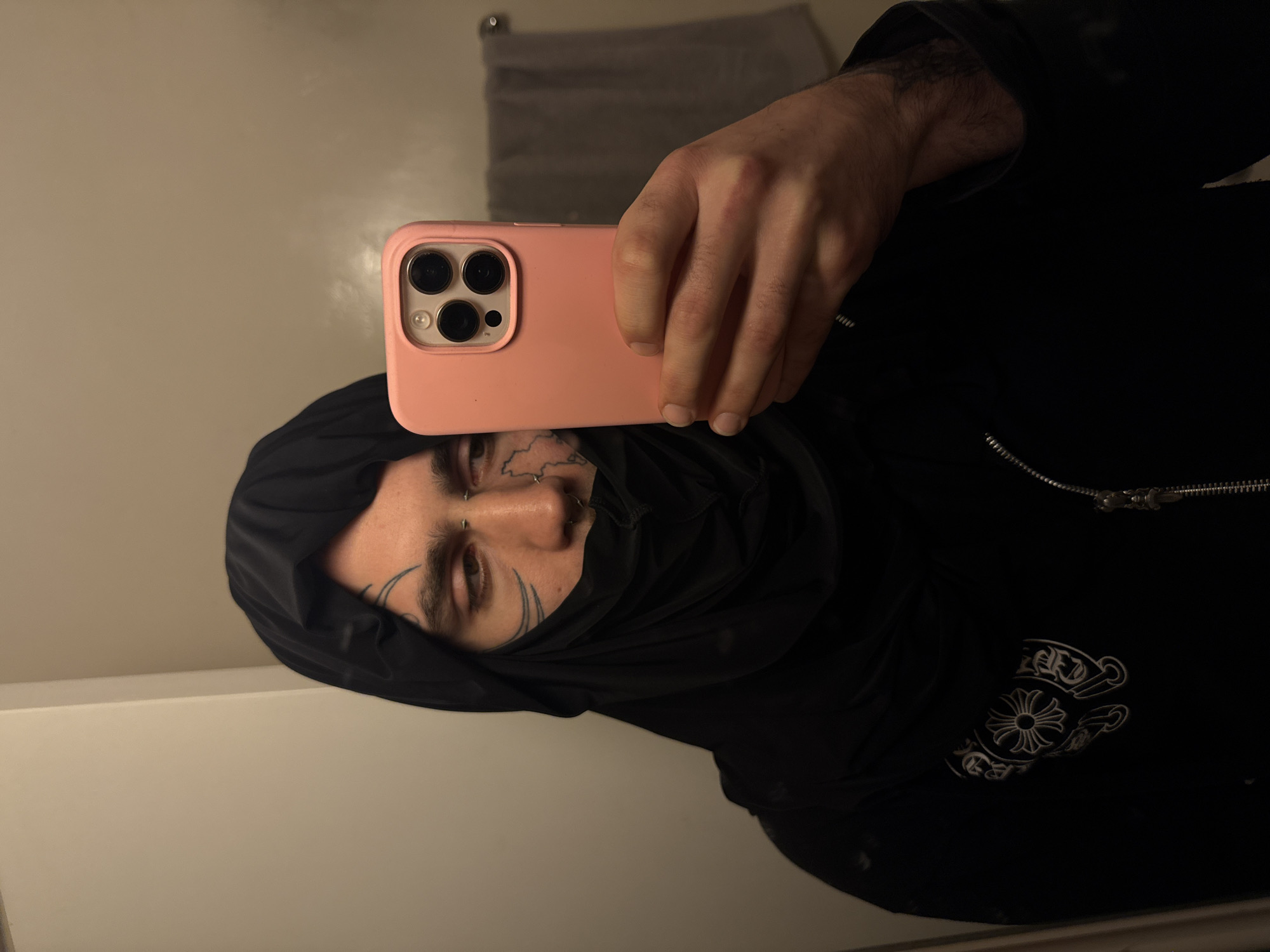 A selfie taken in an indoor setting. The subject is a person with tattoos on their face and wearing a black hooded garment and a black jacket with a prominent white logo. The person is holding a pink smartphone to take the picture. The background includes a light-colored wall and a towel rack with a gray towel. There is no discernible evidence to suggest the picture was taken in the city of Buenos Aires, Argentina.