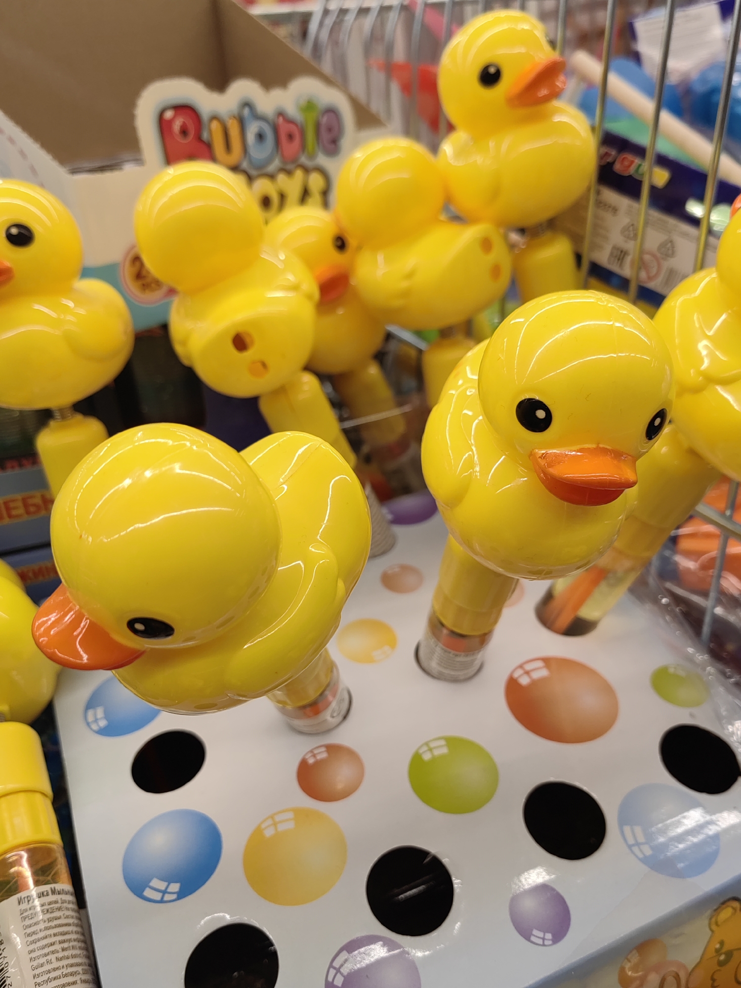 The image is a close-up, eye-level shot of several yellow rubber duck toys arranged on a white display stand. The ducks have orange beaks and black eyes. Some of the ducks are attached to clear plastic tubes, likely bubble blowers, which contain liquid. The display stand has colorful, circular indentations, some of which contain colored shapes resembling eggs, including blue, orange, green, and purple. In the background, there are shelves with more toys and a cardboard display box with "Bubble Toys" written on it. The lighting is bright, suggesting an indoor retail environment. The overall scene depicts a toy store display.