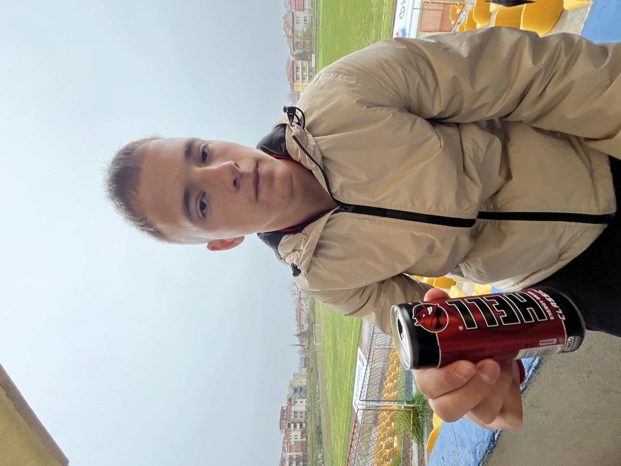 The image depicts a person holding a can of "HELL" energy drink, seemingly at a stadium in Asenovgrad, Bulgaria. The stadium features yellow seating and a green playing field, beyond which are residential buildings. The person is wearing a beige puffer jacket. The weather appears overcast.