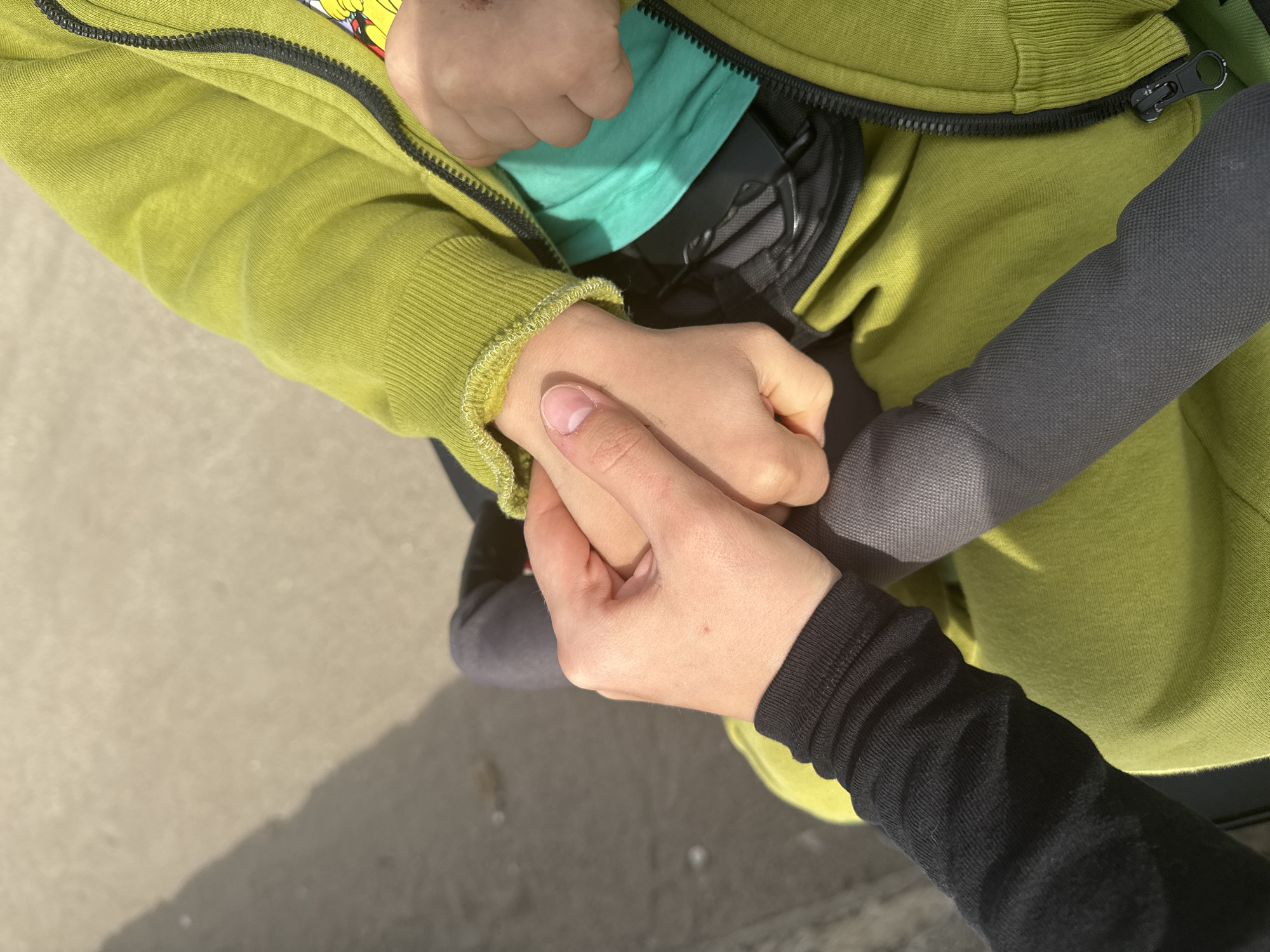 This close-up image captures a tender moment between two individuals, focusing on their hands. One person, likely a child, is wearing a vibrant lime green hooded jacket with a black zipper and a light turquoise t-shirt underneath. Their small hand is gently held by the larger, more mature hand of another person, who is wearing a black long-sleeved top with ribbed cuffs.
The smaller individual appears to be seated, possibly in a stroller or car seat, as parts of a black harness or seat are visible beneath their jacket. A black, textured handle or bar, consistent with a stroller or similar equipment, is also visible on the right side of the frame. The hands are intertwined, conveying a sense of care, comfort, and connection.
The setting is outdoors, characterized by strong, angled shadows cast on a light-colored ground surface, likely concrete or pavement, indicating a sunny day, possibly during the morning or late afternoon. The scene is devoid of any specific location identifiers, though the provided context is Bar, Montenegro. No text is visible in the image.