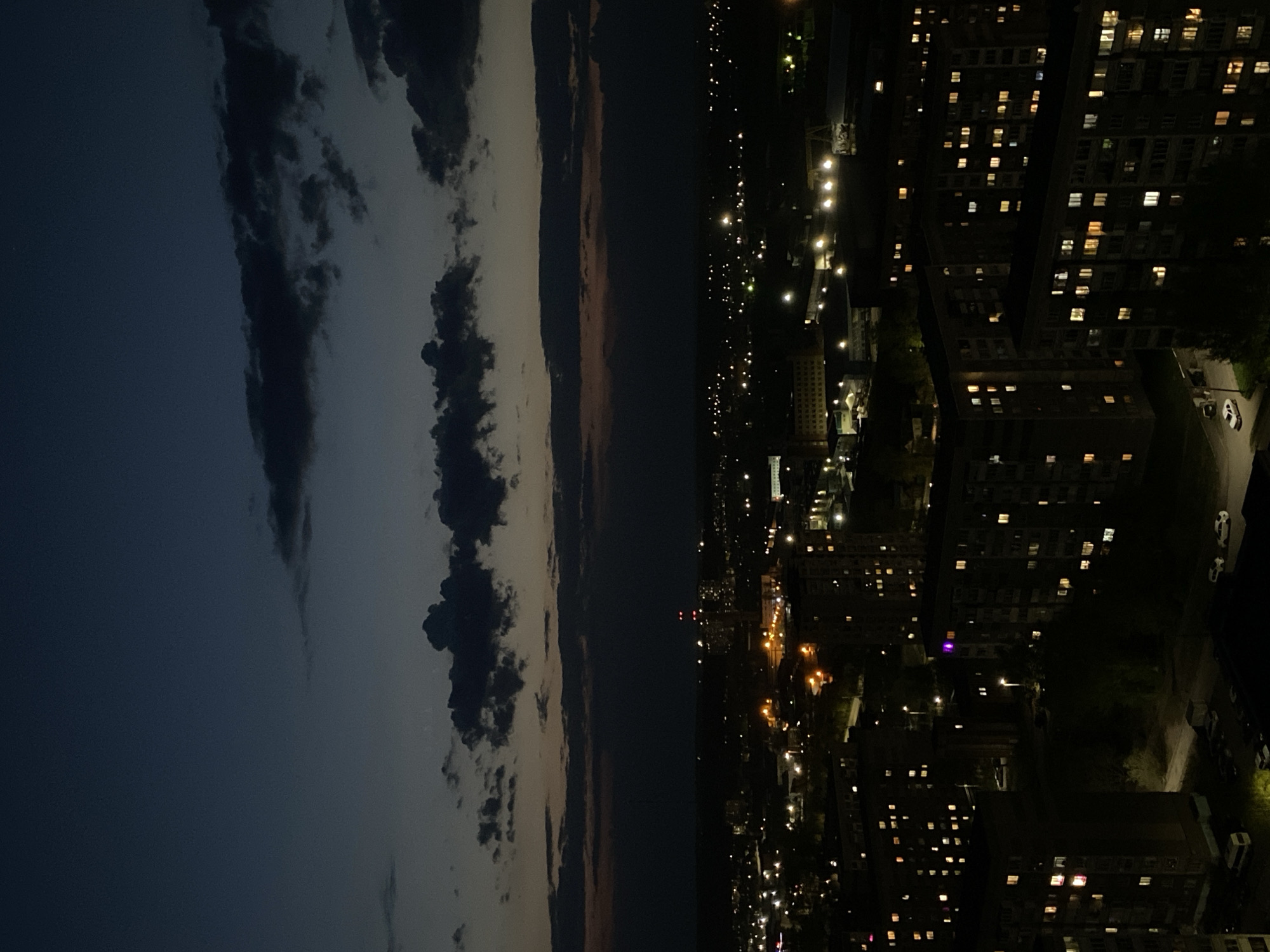 The image is an outdoor night shot depicting a division between a sky with clouds and a cityscape. On the left side, the sky transitions from a deep blue to lighter shades with prominent dark clouds clustered together. On the right, the cityscape is filled with numerous buildings, most of which appear to be residential due to the presence of many illuminated windows. Streetlights and the general glow of the city contribute to the light. There is a road with cars parked along it near some of the buildings. The precise location in Izhevsk, Russia, cannot be determined with certainty from this image alone. The picture appears to be taken from an elevated viewpoint.