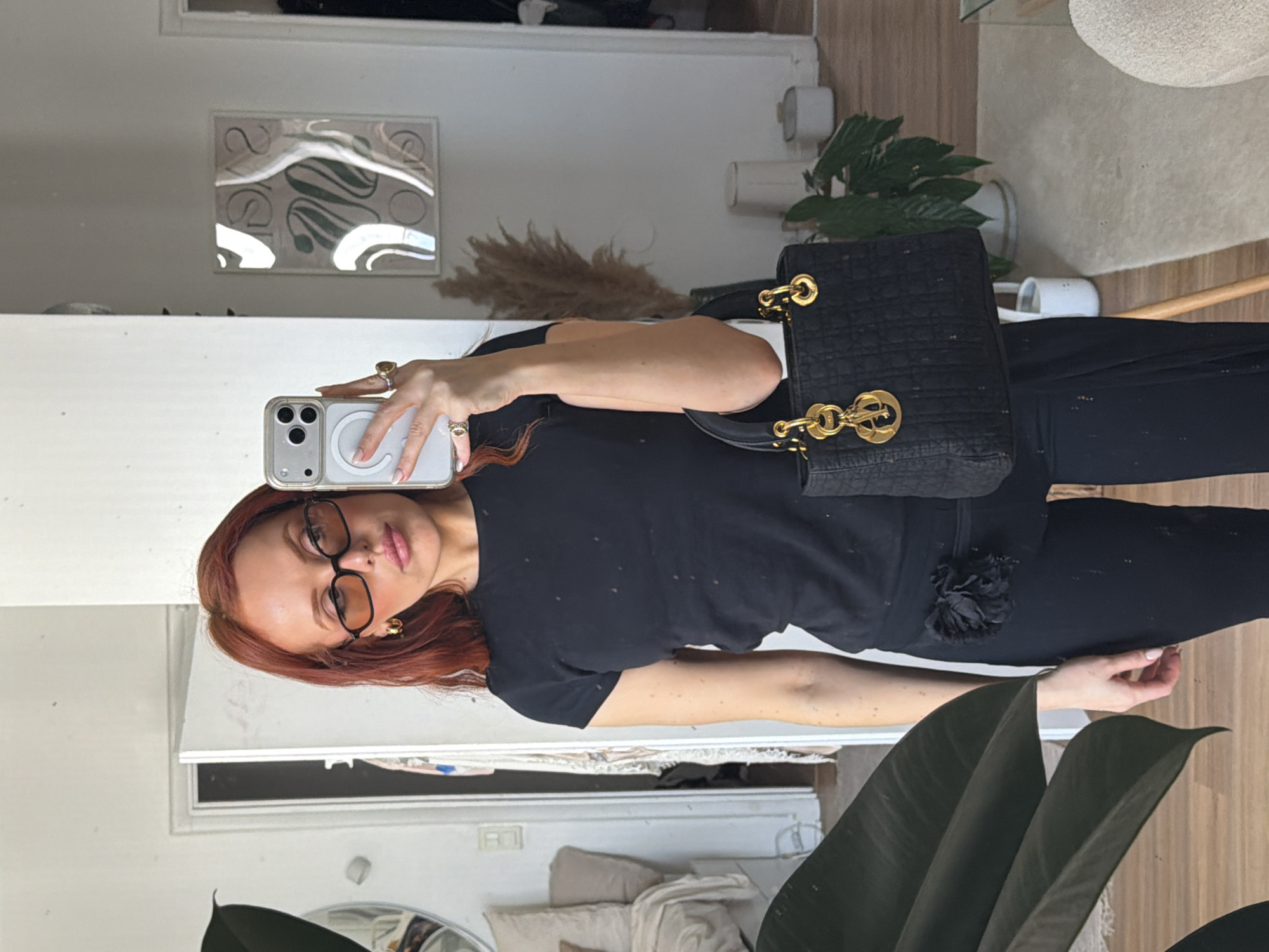 A woman with red hair and glasses is taking a selfie in a mirror. She is holding a black purse and wearing a black outfit. The background includes a plant, a painting, and furniture, suggesting an indoor environment, potentially her home. There is no discernible information to confirm that the scene is in Paris, France.