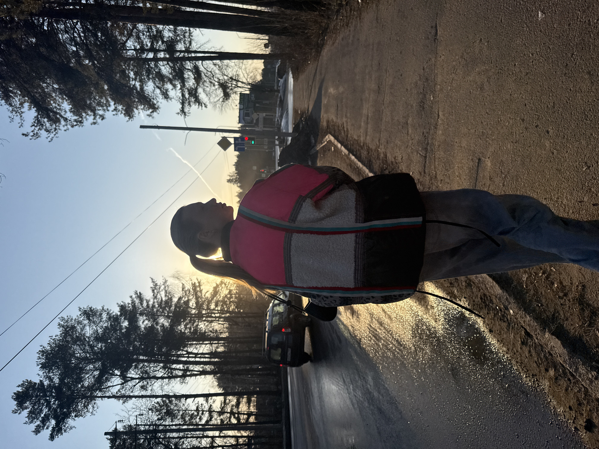 A young person stands on a wet asphalt surface, likely a road or sidewalk, in what appears to be a semi-urban or suburban environment. The individual is facing away from the viewer, wearing jeans and a colorful, patterned jacket with a ponytail hairstyle. A black object, possibly a bag or satchel, is slung over their shoulder. The light suggests it is either sunrise or sunset. In the background, trees line the road, and there is a vehicle in motion. A traffic light is visible in the distance, showing a green light. The location is likely the city of Chaikovsky, Russia.