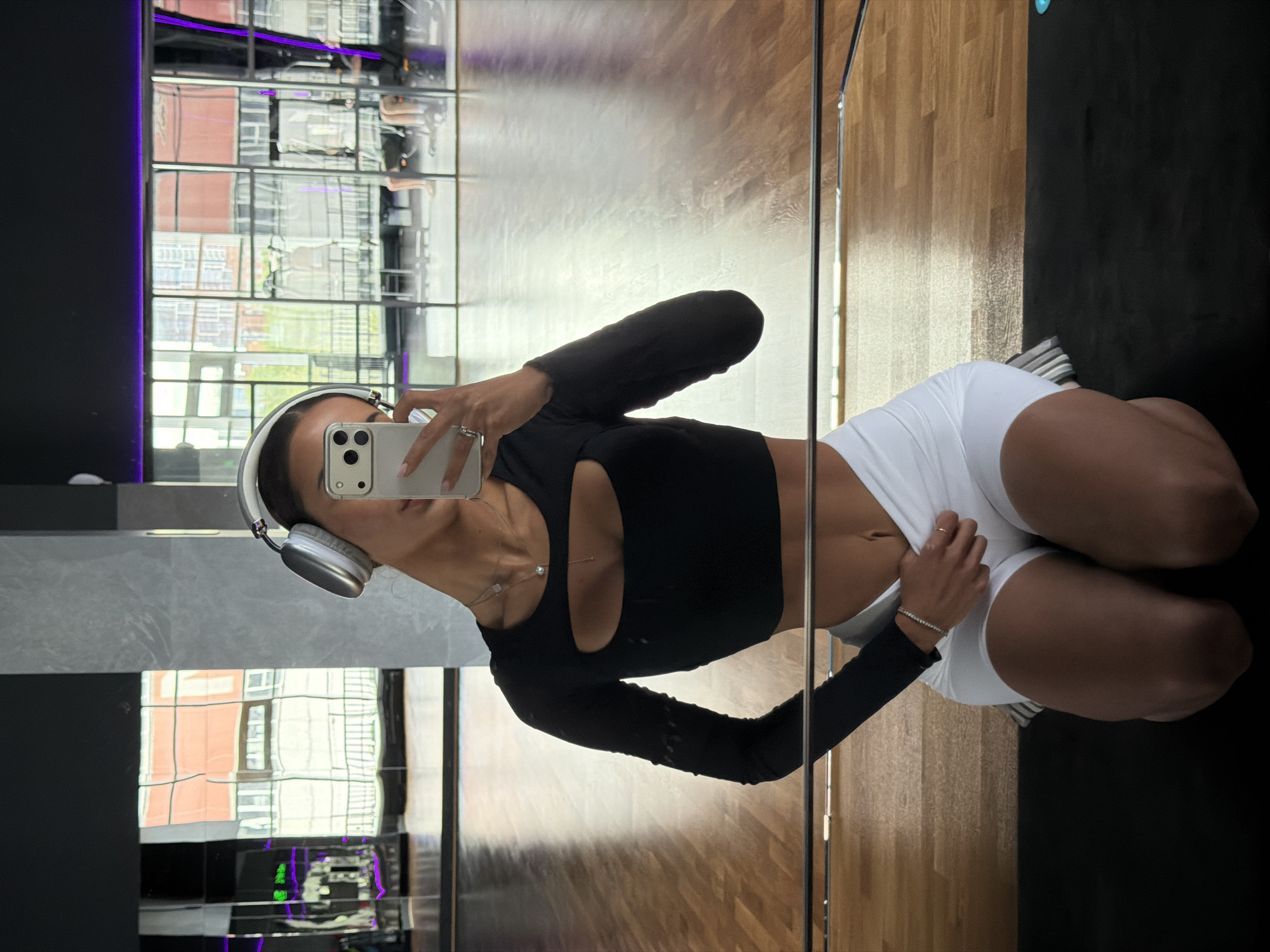 A woman is taking a selfie in a gym. She is kneeling on a black mat in front of a mirror, wearing white shorts, a black long-sleeved crop top with a cutout, and headphones. The gym has wooden floors and a mirrored wall. The reflection in the mirror shows a building with multiple windows, possibly an apartment building or office building. The image was taken in Krasnoyarsk, Russia.
