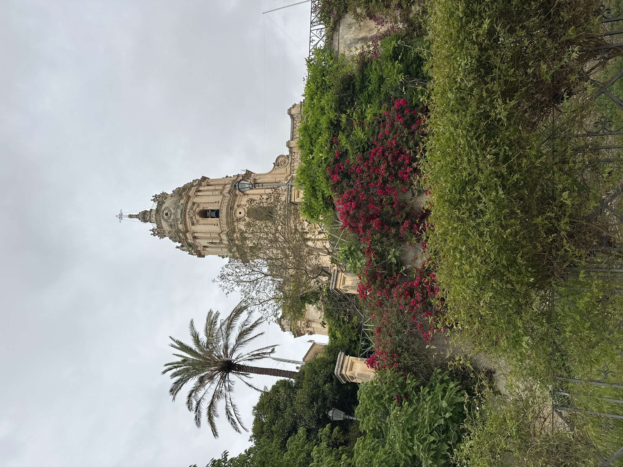 The image shows the facade and dome of the Cathedral of San Giorgio, located in Modica, Italy. Lush greenery and flowering bougainvillea drape over retaining walls and structures adjacent to the cathedral. A palm tree stands in the foreground. The sky is overcast.