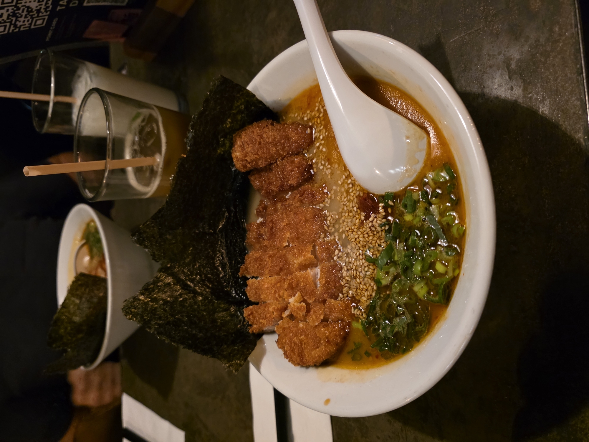 A close-up shot captures a bowl of ramen situated on a dark table, likely indoors. The ramen bowl features a white ceramic spoon, fried cutlets of breaded meat, dried seaweed, sesame seeds, and chopped green onions. A second, smaller bowl of ramen is partially visible in the background. Beside the bowls, there are two glasses, each containing a straw.
The image was taken in Ensenada, Mexico.