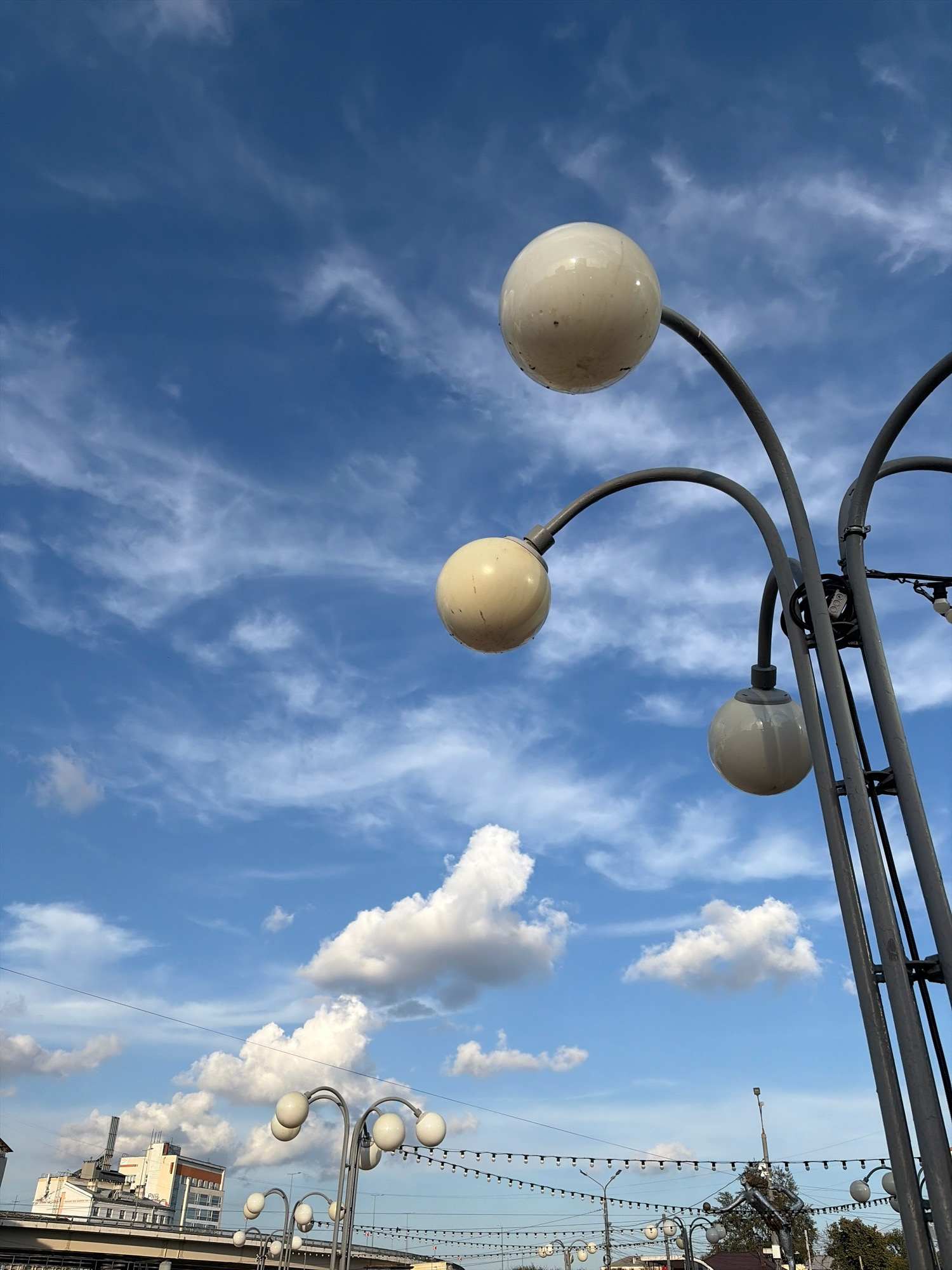 The image shows a view of the Nizhny Novgorod embankment against a blue sky with scattered clouds. In the foreground, there is a tall lamppost with multiple spherical lights. Further back, there are more lampposts and strings of decorative lights. In the background, there are buildings, some of which appear to be industrial or commercial, and trees. The scene is viewed from a slightly elevated perspective, looking across the embankment.