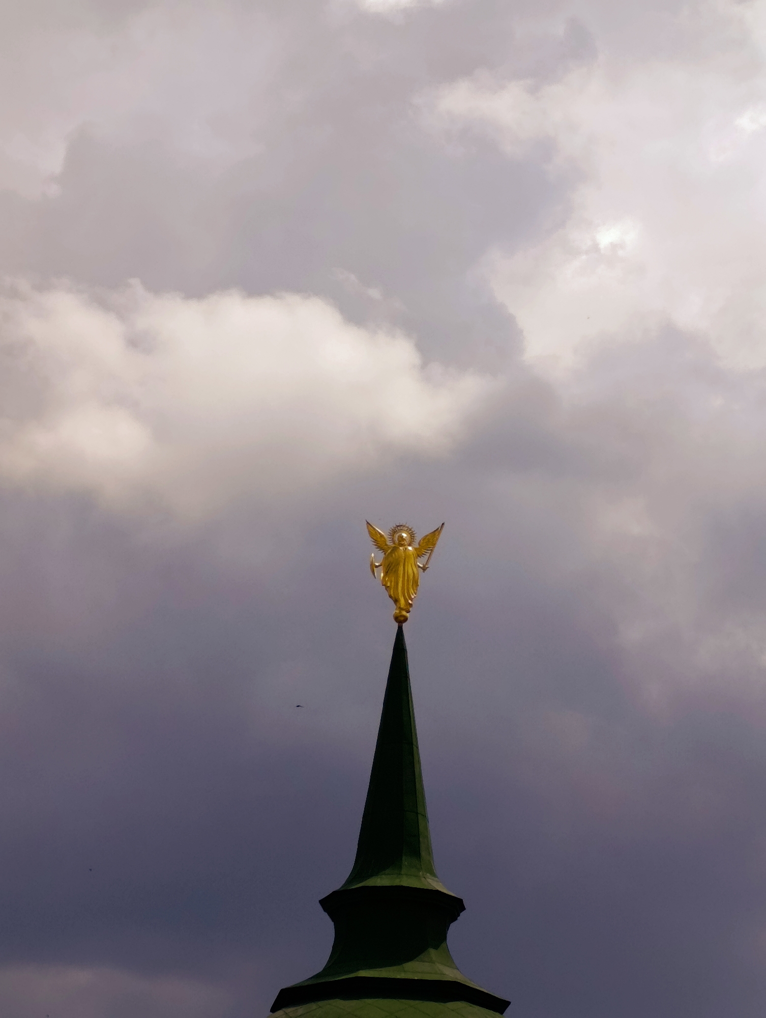 This is a low-angle, close-up shot of the golden angel statue atop the spire of St. Nicholas Cathedral in Kyiv, Ukraine. The statue, depicting an angel with outstretched wings and a halo, is the focal point, positioned against a dramatic sky filled with heavy, grey clouds. The spire itself is dark green and pointed, tapering to a sharp point where the angel stands. The overall mood is somber and imposing due to the dark clouds and the ominous lighting.