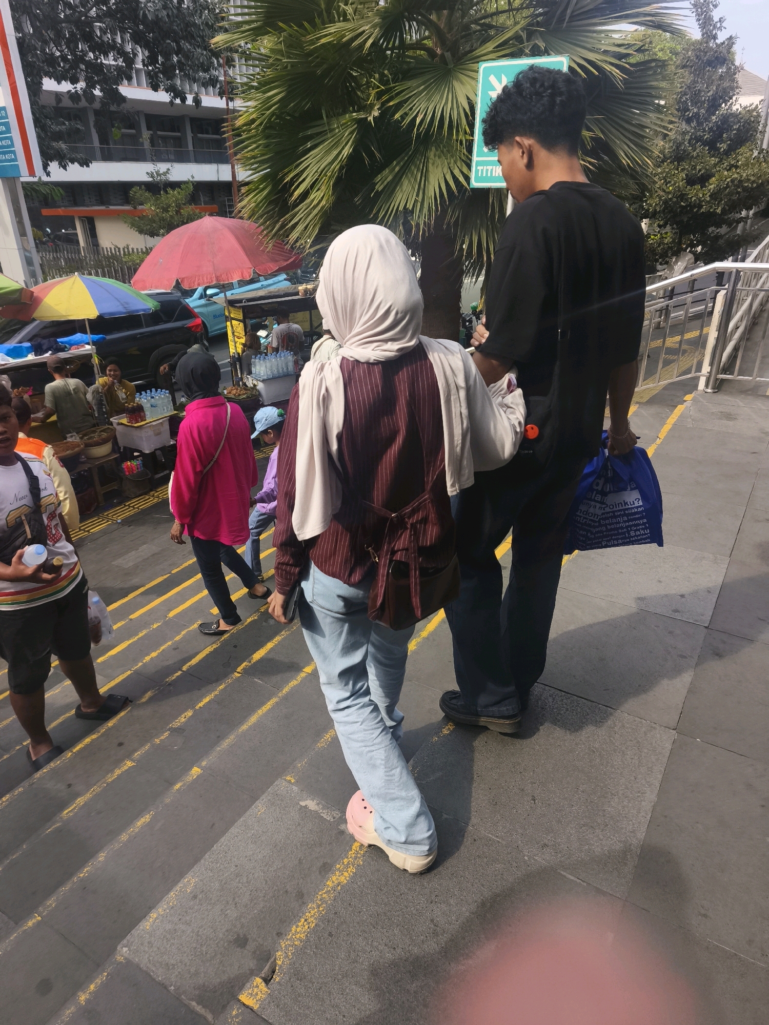 The image depicts an outdoor scene on a sunny day, likely in a city environment. In the foreground, two individuals are walking up a set of grey stone stairs with yellow painted lines marking the edges and some of the risers. The person on the left is wearing light blue jeans, a maroon and white striped vest over a light-colored garment, and a light beige headscarf. They are also wearing pink clogs on their feet. The person on the right is wearing dark blue pants and a black t-shirt. They are carrying a large blue shopping bag in their right hand.
In the background, there are several market stalls with colorful umbrellas, selling various items including drinks and food. A bright red umbrella is prominent, positioned above a blue car. Trees with large fronds, possibly palm trees, are visible, along with buildings and other urban structures. A green sign with white text and an icon is also present, attached to a pole. The overall impression is a bustling street scene with pedestrian activity and commercial vendors. Based on the available visual cues, the location is identified as West Jakarta, Indonesia.