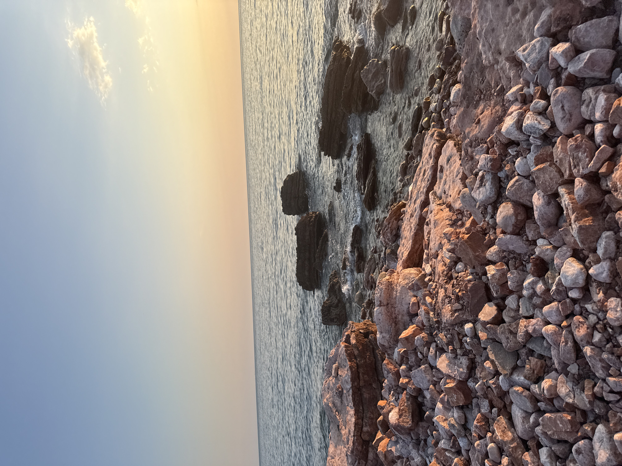 The image depicts a serene coastal scene at sunset in Kamenovo, Montenegro. The foreground is dominated by a rocky shoreline, with numerous stones of varying sizes and shades of grey and pink scattered across the ground. The rocks are rough and uneven, hinting at a natural, untamed landscape.
The sea stretches out to the left of the frame, its surface rippling gently under the soft light. The water is a deep blue, reflecting the warm hues of the sky. Further out, the horizon is a soft gradient of colors, transitioning from a pale blue near the water to a warm, golden orange as it meets the sky. A few wispy clouds are visible in the upper left portion of the image, catching the last rays of the setting sun.
The lighting suggests that the photograph was taken during the golden hour, as the low sun casts a warm, reddish glow on the rocks and creates a tranquil atmosphere. There are no people or man-made objects visible, emphasizing the natural beauty of the location. The overall mood is peaceful and contemplative, evoking a sense of calm and solitude. The scene is a quintessential representation of a Mediterranean sunset by the sea.