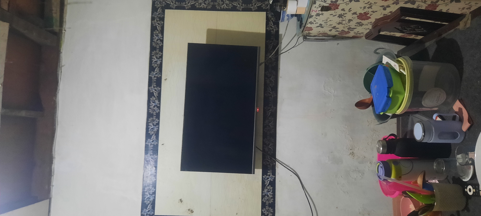 The image depicts an indoor scene, likely a room in a residential setting in North Jakarta, Indonesia. A flat-screen television is mounted on a wall, with a decorative border around it. Below the television, a table holds various items, including water bottles, cups, a bucket, and food containers. A portion of a patterned cabinet and a wooden chair are also visible. The wall appears to have some imperfections, possibly water stains or peeling paint. The lighting suggests it is either evening or the room is dimly lit. There are no people visible in the image.