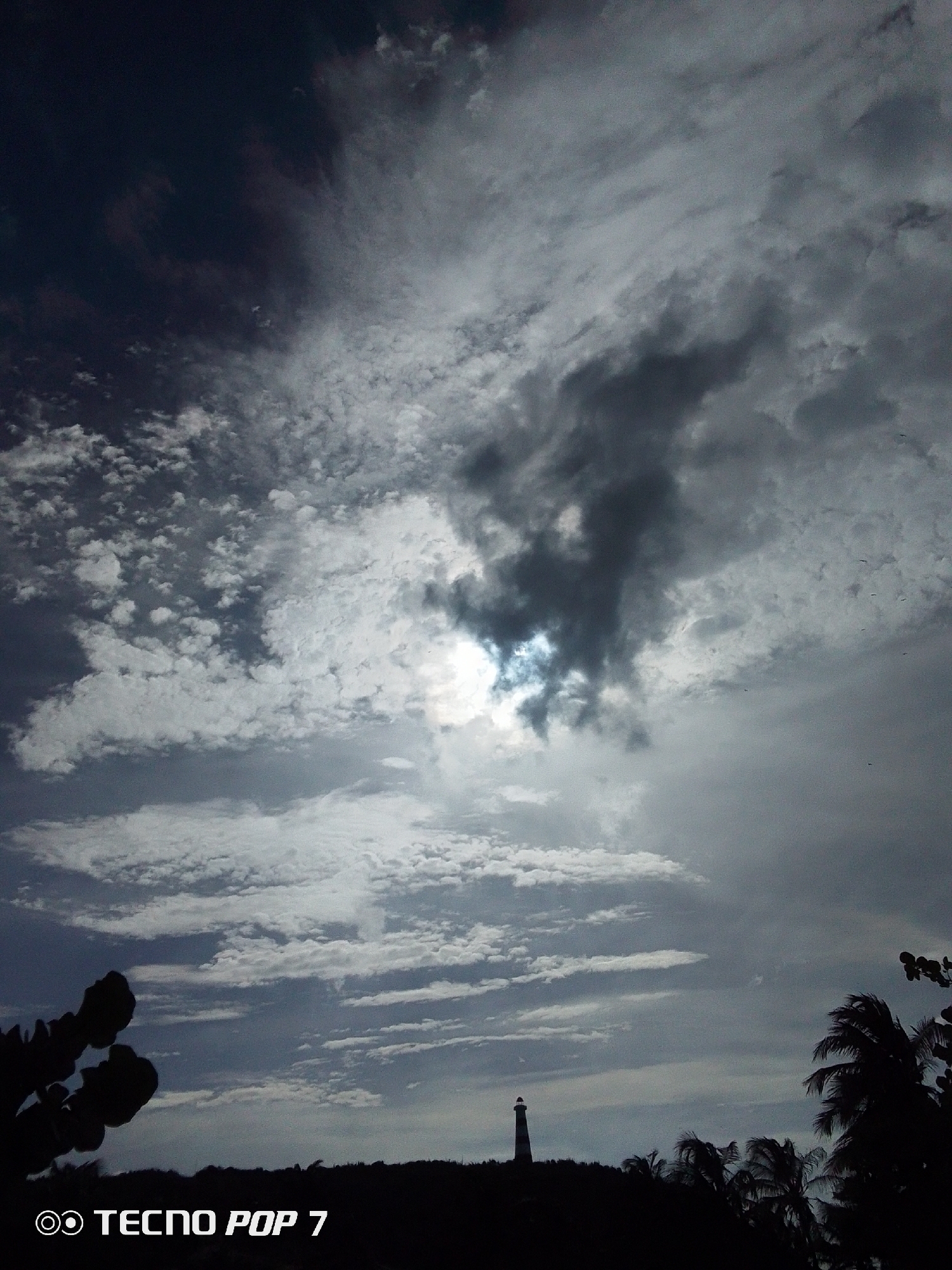 The image captures a monochrome scene of a cloudy sky with a silhouette of a landscape below, likely in Pedro González, Venezuela. A lighthouse stands out on the horizon. The sky is filled with varied cloud formations, with a darker, dense cloud partially obscuring the sun. The foreground includes silhouetted vegetation, including what appears to be cacti on the left and palm trees on the right, indicating a tropical or coastal environment. The bottom left corner displays a "TECNO POP 7" watermark.