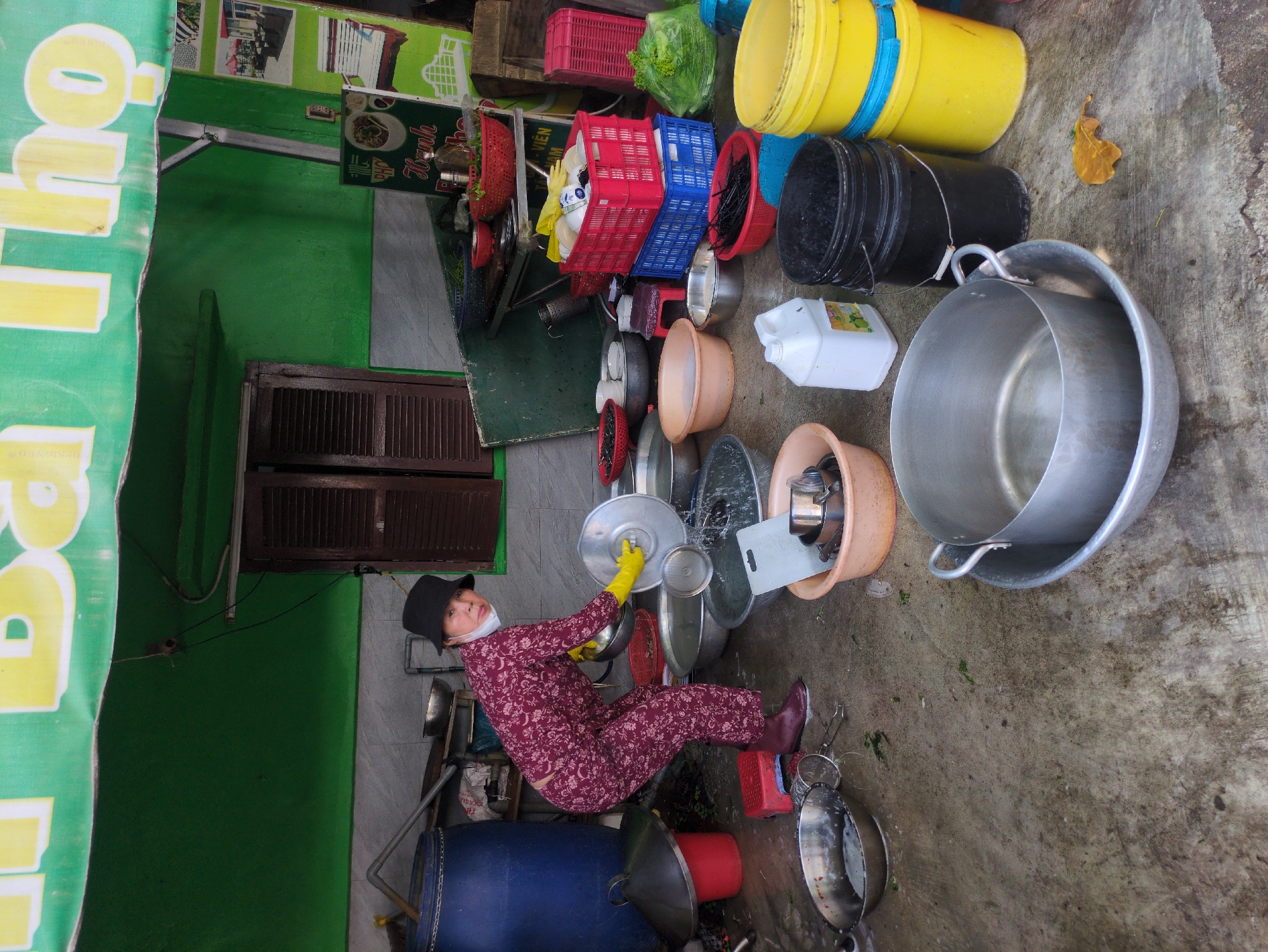 A woman wearing a patterned maroon jumpsuit, black hat, face mask, and yellow gloves is washing dishes outside in Tuy Hòa, Vietnam. She is sitting surrounded by various sized metal bowls and pots, and a blue plastic barrel is behind her. Baskets and buckets are scattered nearby. The setting is adjacent to a green wall with signage in Vietnamese. The ground is concrete.
