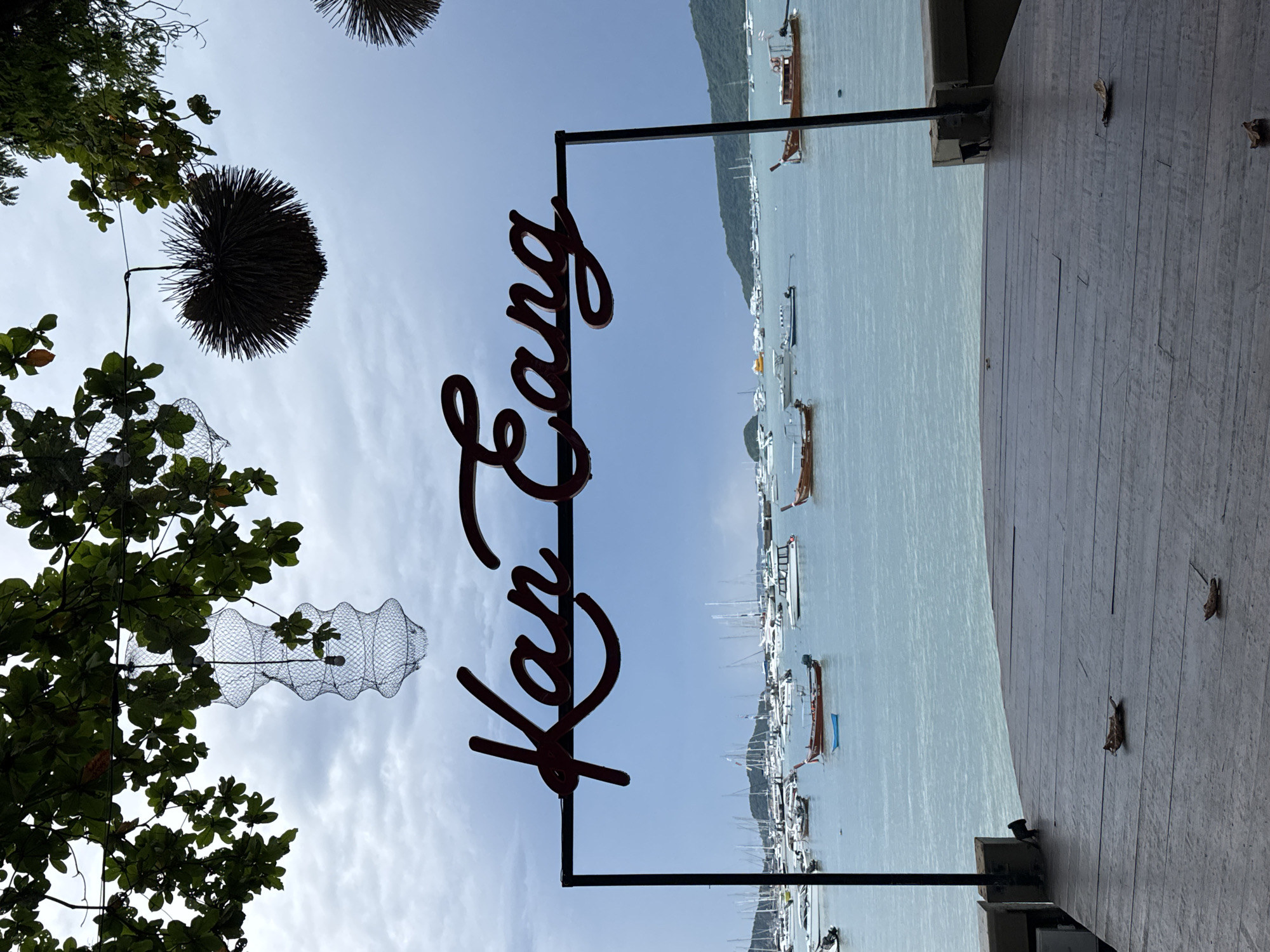 A scenic view of a harbor in Rawai, Thailand, is presented. In the foreground, a wooden deck with a slight curve leads the eye towards the water. The deck is weathered, with visible wood grain and a few scattered leaves. A prominent dark metal frame supports a stylized, dark red cursive sign that reads "Kan Eang." Beyond the sign, the tranquil blue water of the harbor is dotted with numerous boats of various sizes, including sailboats and smaller wooden vessels. In the background, a lush green hillside rises, partially obscured by a cloudy sky. Overhead, the branches of a tree, adorned with decorative string lights and a dark, spiky spherical ornament, frame the upper portion of the image, adding a touch of ambiance to the waterfront scene. The overall atmosphere is peaceful and inviting, capturing the essence of a seaside establishment.