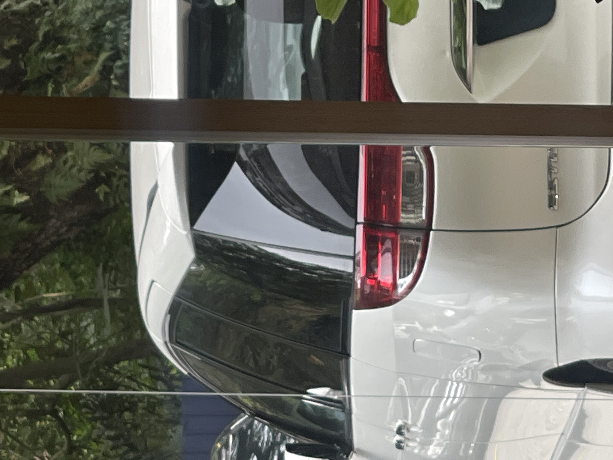 The image captures the rear-left side of a white minivan, possibly parked outdoors. The vehicle has tinted windows and visible red taillights. A brown wooden pillar partially obstructs the view of the vehicle. In the background, lush green foliage and trees are present, suggesting a natural or park-like setting. The environment appears to be an outdoor space in Kuala Lumpur, Malaysia, given the presence of tropical vegetation. The lighting indicates it is daytime.