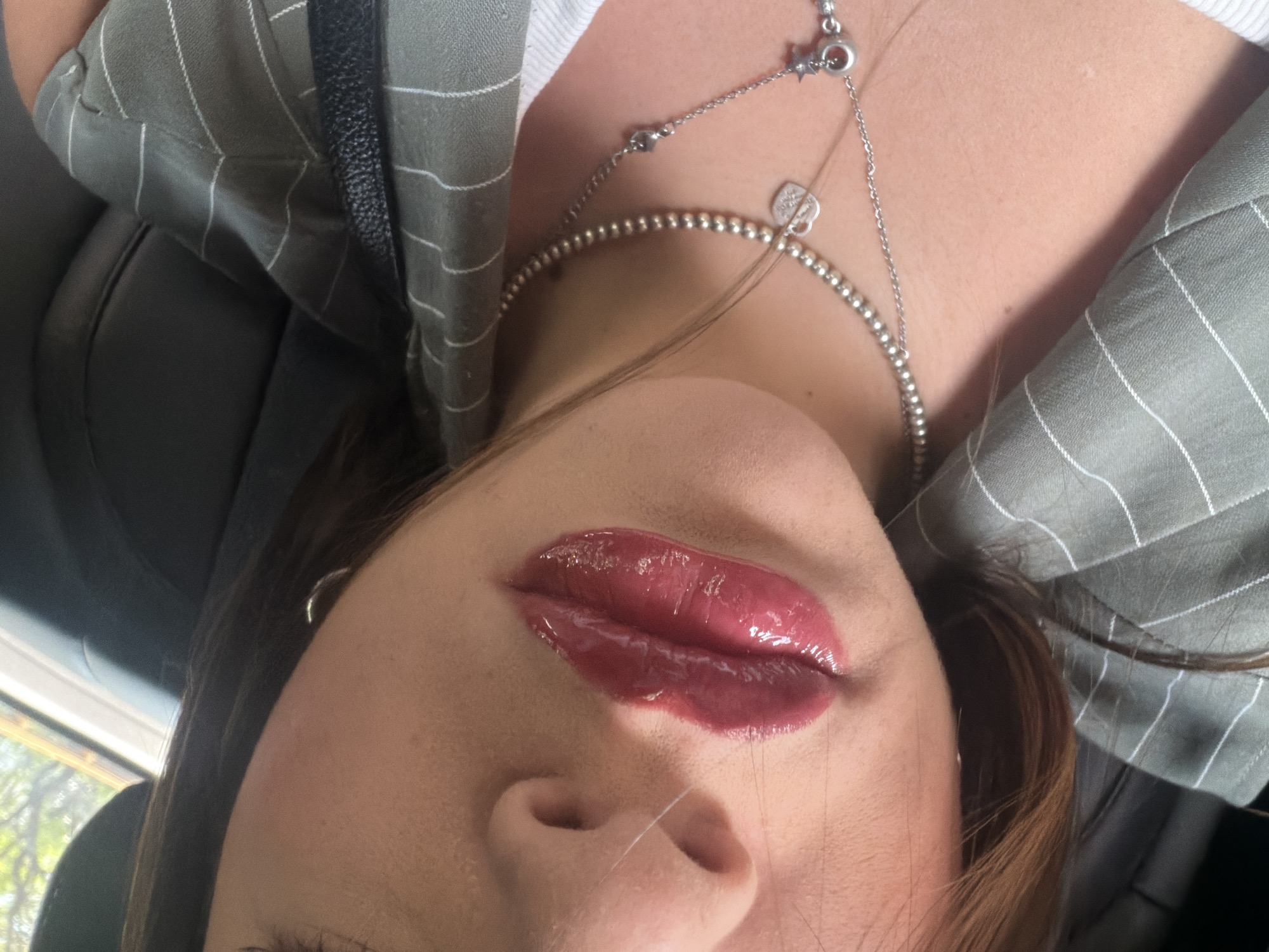 A close-up, angled selfie depicts a woman from the chest up. She wears a gray, pinstriped blazer and a white top underneath. Two silver necklaces adorn her neck. The focal point is her face, particularly her lips, which are coated in a glossy, deep red lipstick. The image was taken in a vehicle, with a glimpse of foliage visible through the window in the background, which points towards the presence of greenery in an urban setting, possibly Mexico City. The interior of the vehicle is dark.