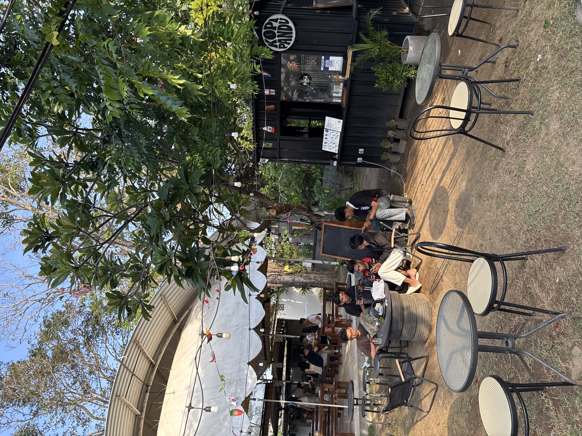 This image depicts an outdoor seating area at a coffee shop, possibly "Kopi Pandu," in Denpasar, Indonesia. The setting is informal and rustic, with a mixture of seating arrangements and natural elements. Several groups of young men are seated at tables, engaged in conversation or looking at their phones. The tables are made of various materials, including what appears to be stacked tires for one larger table. Chairs are a mix of styles, some made of metal and others appearing to be foldable.
The coffee shop itself is housed in a black, modular structure, resembling a shipping container, with a service counter and windows. Decorative flags from different countries are strung above the seating area and the coffee shop. String lights are also present, suggesting the possibility of evening operation. Lush green trees and foliage surround the area, creating a shaded and natural ambiance. The ground is a mixture of dirt and grass, with fallen leaves scattered around. Sunlight casts shadows, indicating it is likely late afternoon. In the background, there is a covered seating area with more tables and patrons. The overall scene suggests a relaxed and casual atmosphere for patrons to enjoy coffee and socializing outdoors.