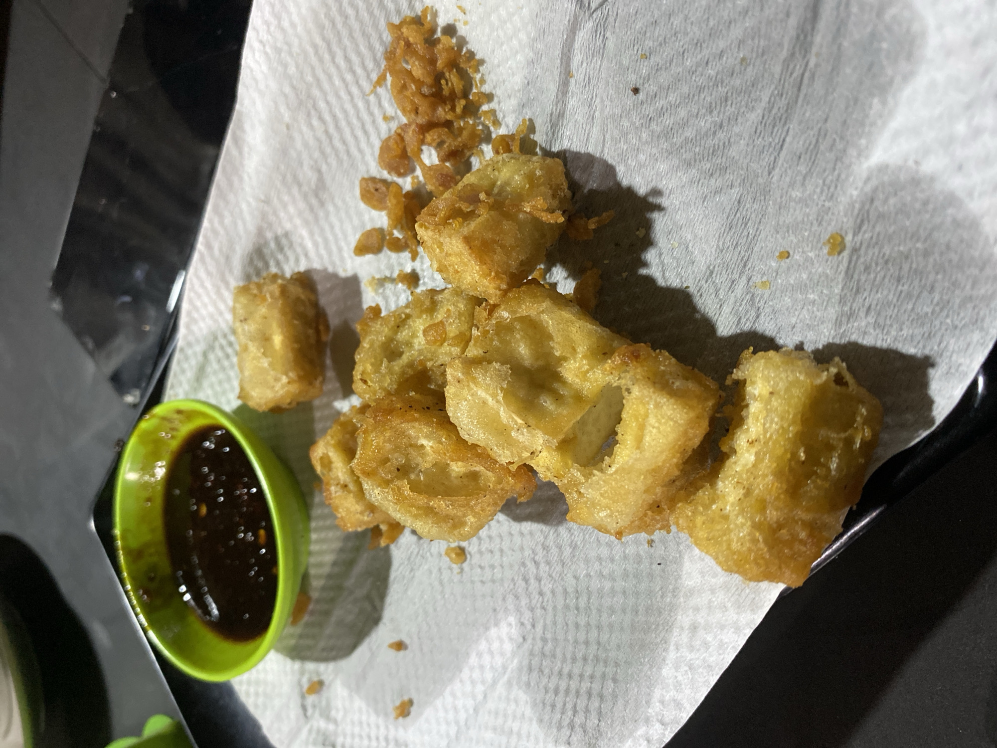 The image shows a close-up of fried tofu on a white paper towel. A small green bowl containing a dark sauce sits to the left of the tofu. The tofu pieces are irregularly shaped and golden-brown in color, with some scattered crispy fried bits nearby. The paper towel rests on a dark surface, possibly a tray or table. The location is Gampongaceh, Indonesia. There are no people visible in the image.