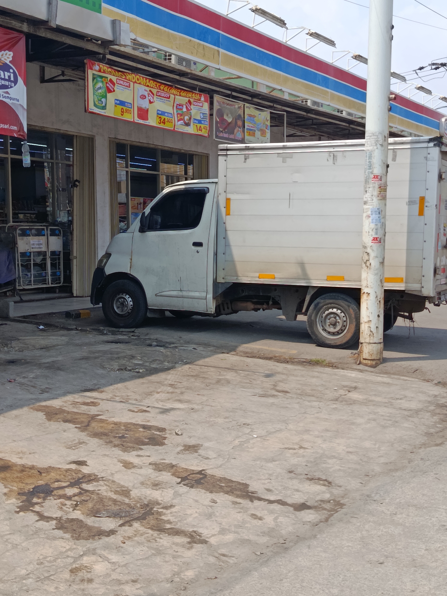 In Wanasari, Indonesia, a white box truck is parked on a paved area in front of a store. The truck is facing right, with its rear cargo box covering most of the vehicle's length. The truck's tires are visible, as is the driver's side door and window. In the background, the storefront of a convenience store is visible, with awnings and advertisements displayed. A white pole stands to the right of the truck, covered in various posters and peeling paint. The ground in front of the store is concrete and shows signs of wear and tear, with some staining and debris scattered around. The overall scene depicts a typical urban commercial setting in Indonesia, with a focus on the parked truck and the adjacent store.