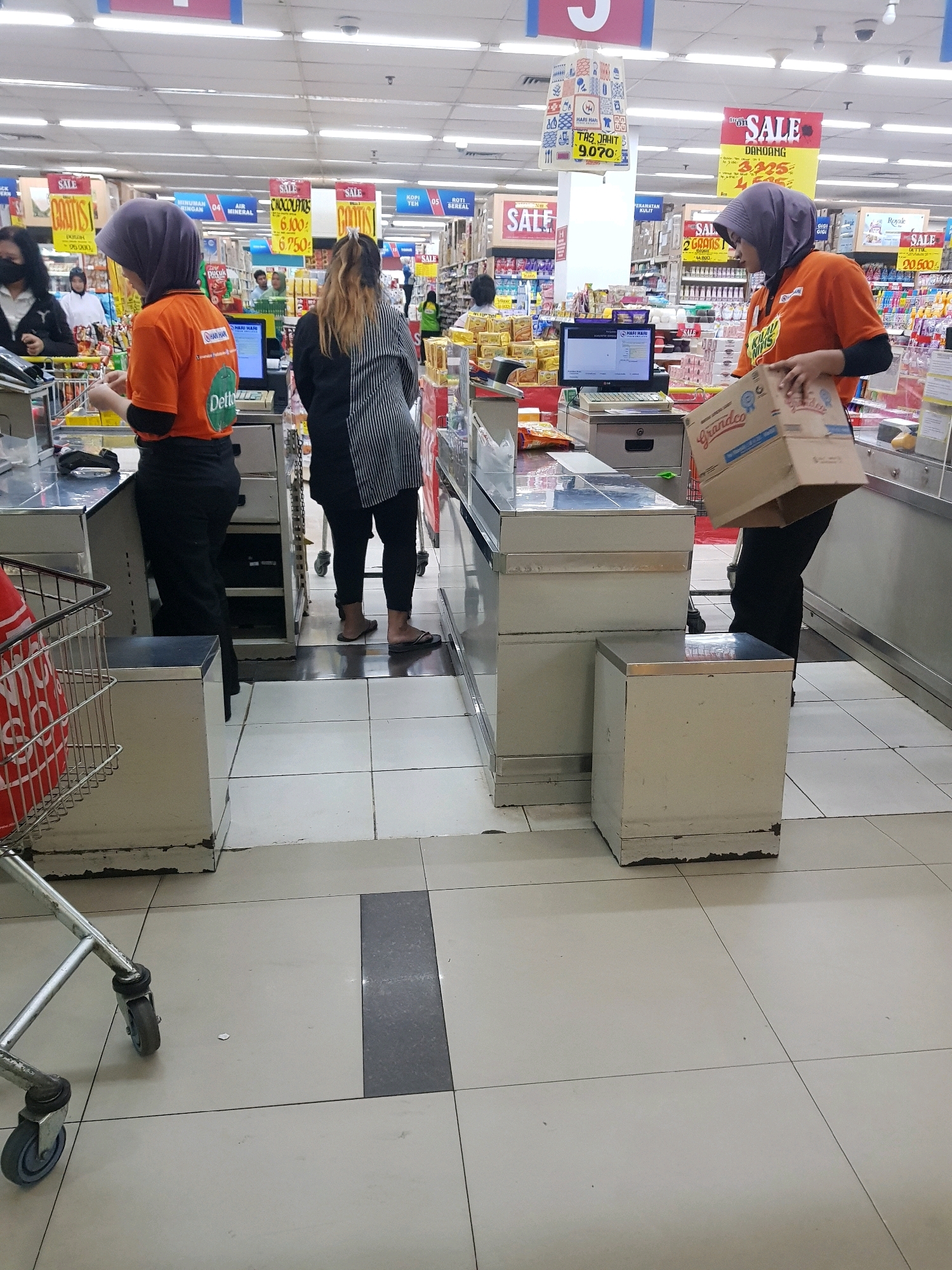 In a grocery store in Bekasi, Indonesia, two cashiers and one customer are visible at the checkout counters. A cashier in an orange shirt, wearing a purple hijab, stands at a counter. To her right, another cashier, also in an orange shirt and purple hijab, is holding a cardboard box labeled "Grandco". A customer with long blonde and brown hair, wearing a black and white striped shirt, stands between the two cashiers. The store environment features overhead fluorescent lighting and shelves stocked with various products. Red shopping baskets and a shopping cart are also present. The floor is tiled in a grid pattern.
