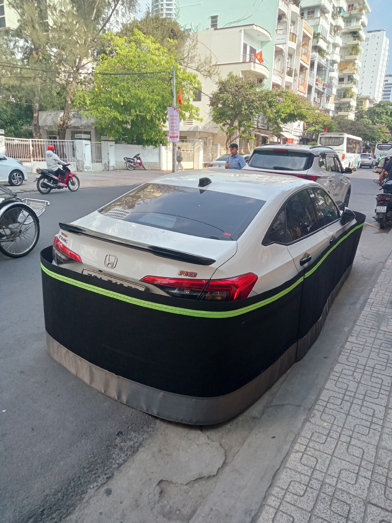 A white Honda Civic RS sedan, distinguished by a unique black, skirt-like protective cover with a bright green stripe, is parked on the side of a bustling street in Nha Trang, Vietnam. The custom cover extends from the car's lower body panels to the ground, likely designed to shield against scratches or minor impacts in the dense urban environment.
The scene is set on a clear, sunny day, indicated by the bright light and distinct shadows, suggesting mid-morning or early afternoon. Numerous motorbikes are present, including a red one being ridden by a person in a red helmet on the left, and another parked on the right with a partially visible license plate reading "79-V1 556.36," confirming the Khánh Hòa province location. A silver SUV is parked directly behind the Civic, with a man in a blue shirt standing beside it. Further down the street, a white bus is visible.
Multi-story buildings with balconies, characteristic of Vietnamese architecture, line the street, interspersed with vibrant green trees. A paved sidewalk with patterned tiles runs along the right side. A utility pole with a red banner displaying Vietnamese text, including fragments like "CÔNG," "TRƯỜNG," and "ĐÀO TẠO" (training), suggests a public announcement or educational institution. The car itself clearly displays "CIVIC" and "RS" badges, along with the Honda emblem on its rear.