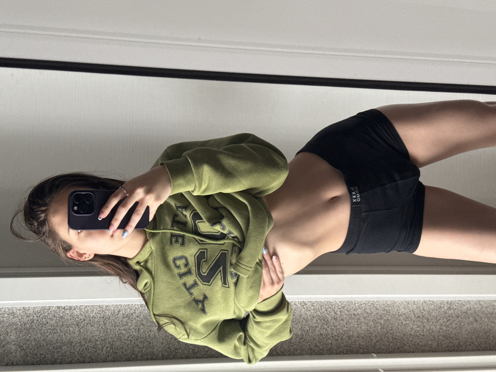 An indoor mirror selfie captures a young person, likely female, taking a photograph with their smartphone. The subject's face is largely obscured by the dark-colored smartphone, revealing only one eye and part of their cheek. They have dark hair and are wearing a small stud earring and a silver ring on their index finger. Their nails are painted, featuring a light color and a design on at least one finger.
The individual is dressed in an olive green hooded sweatshirt (hoodie) with a distressed appearance. Black lettering on the hoodie is partially visible, appearing to spell "STATE CITY," with prominent "S" and "T" characters. Below the hoodie, a toned midriff is visible, extending to a pair of black athletic or spandex shorts. The waistband of the shorts displays mirrored text that reads "OMOX MAXX." One hand holds the phone, while the other rests on their side/hip, suggesting a pose.
The setting is an indoor room, visible through the mirror's reflection, showing a light-colored wall or door and a grey carpeted floor. The lighting is bright, indicating a well-lit interior, possibly during the daytime. This image is associated with Sofia, Bulgaria.