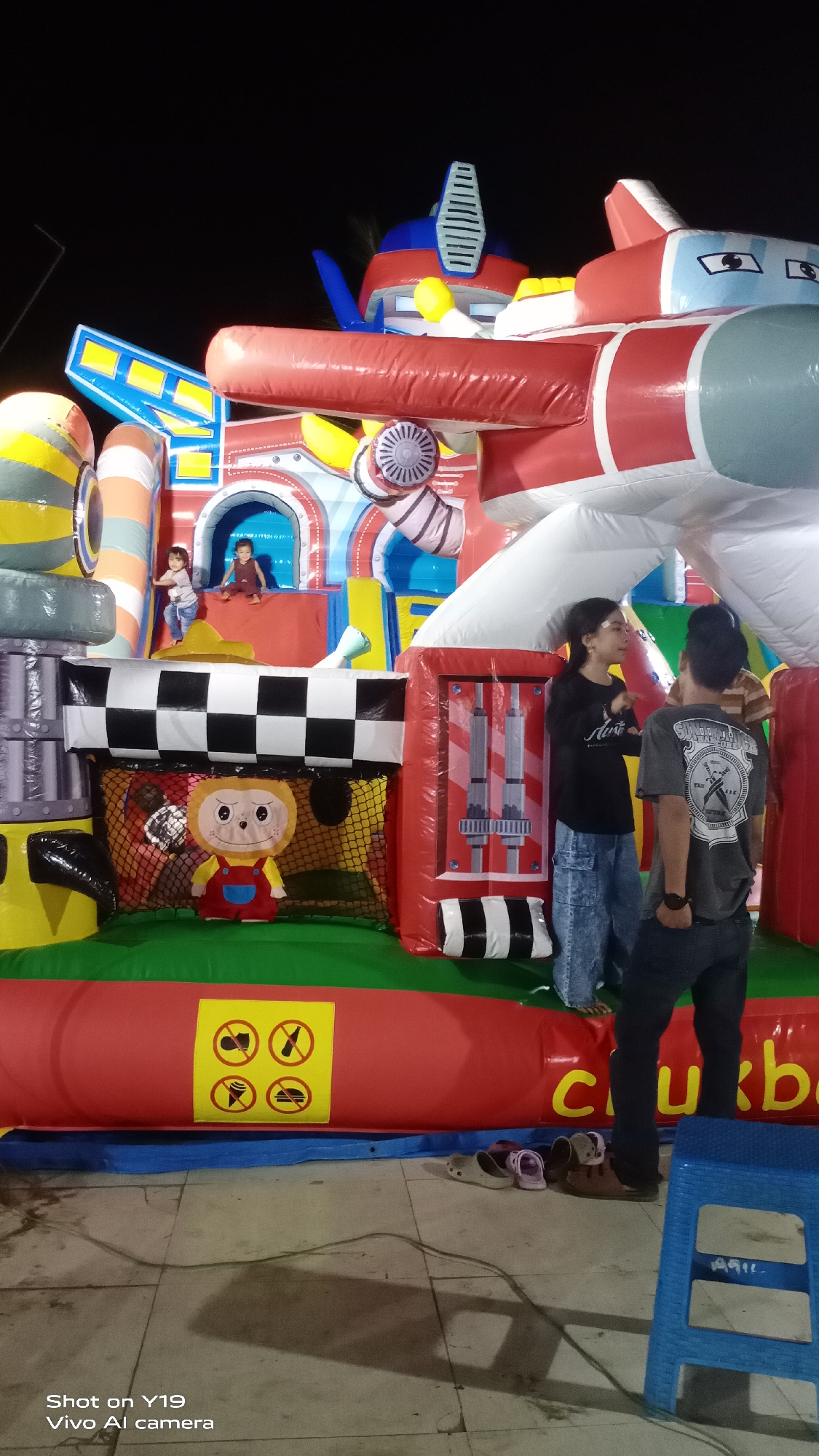 A group of people, including children and adults, are gathered around a large inflatable playground resembling a robot. Two children are visible inside the inflatable structure, one sitting and the other jumping. Two adults are standing outside, engaged in conversation. The playground features a checkered pattern and a plush monkey toy. Yellow warning signs depicting crossed-out food items are posted on the red inflatable base. Several pairs of footwear are scattered on the ground near the playground. The scene appears to be set outdoors at night, with a dark sky in the background. The location is identified as Sea, Indonesia.