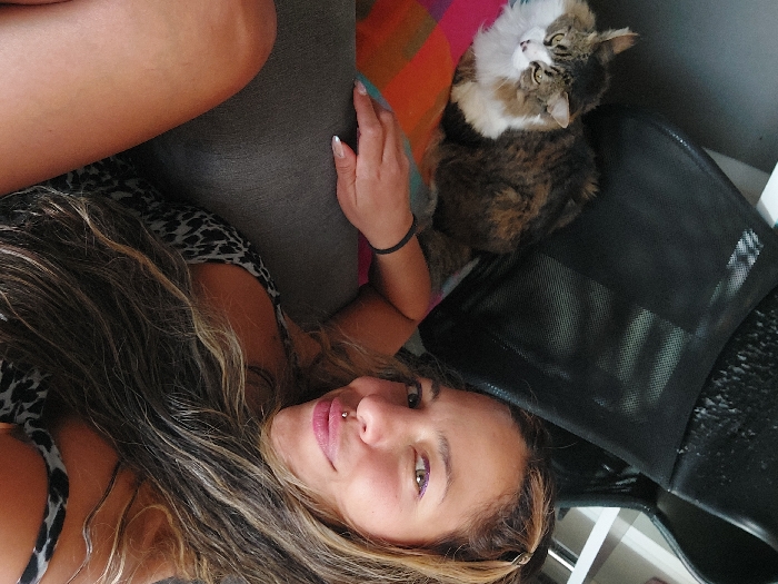 A woman lies sideways on a gray couch. Her hair is long and wavy, with blonde highlights. She wears a black and white patterned top. A cat sits near her, on a black mesh chair. The cat is brown, white, and tan, looking upward. A colorful blanket is seen near the cat. The woman looks at the camera with pink lips and a piercing above her upper lip. A black bracelet is on her wrist.