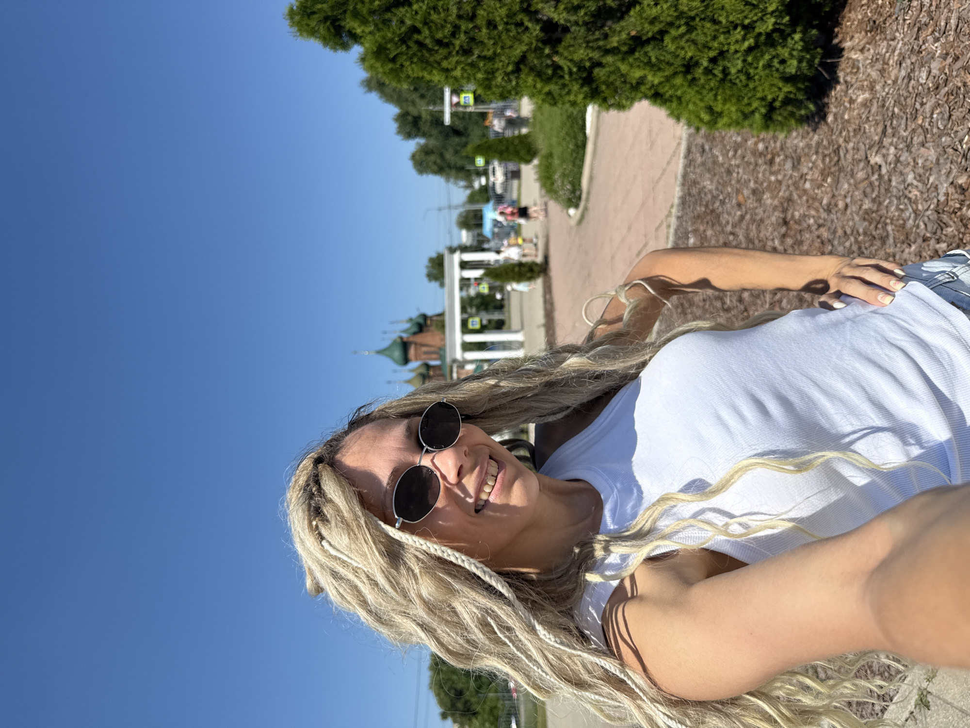 The image is a selfie taken by a woman with blonde dreadlocks and sunglasses. She is wearing a white tank top and denim shorts. She is smiling and looking at the camera. The background shows a clear blue sky, trees, and a building with green domes, which appears to be a church. There are also people walking in the background. The location is the city of Zavolzhye, Russia. The foreground is dominated by the woman and her hair, with the background slightly out of focus. The lighting suggests it is daytime and sunny.