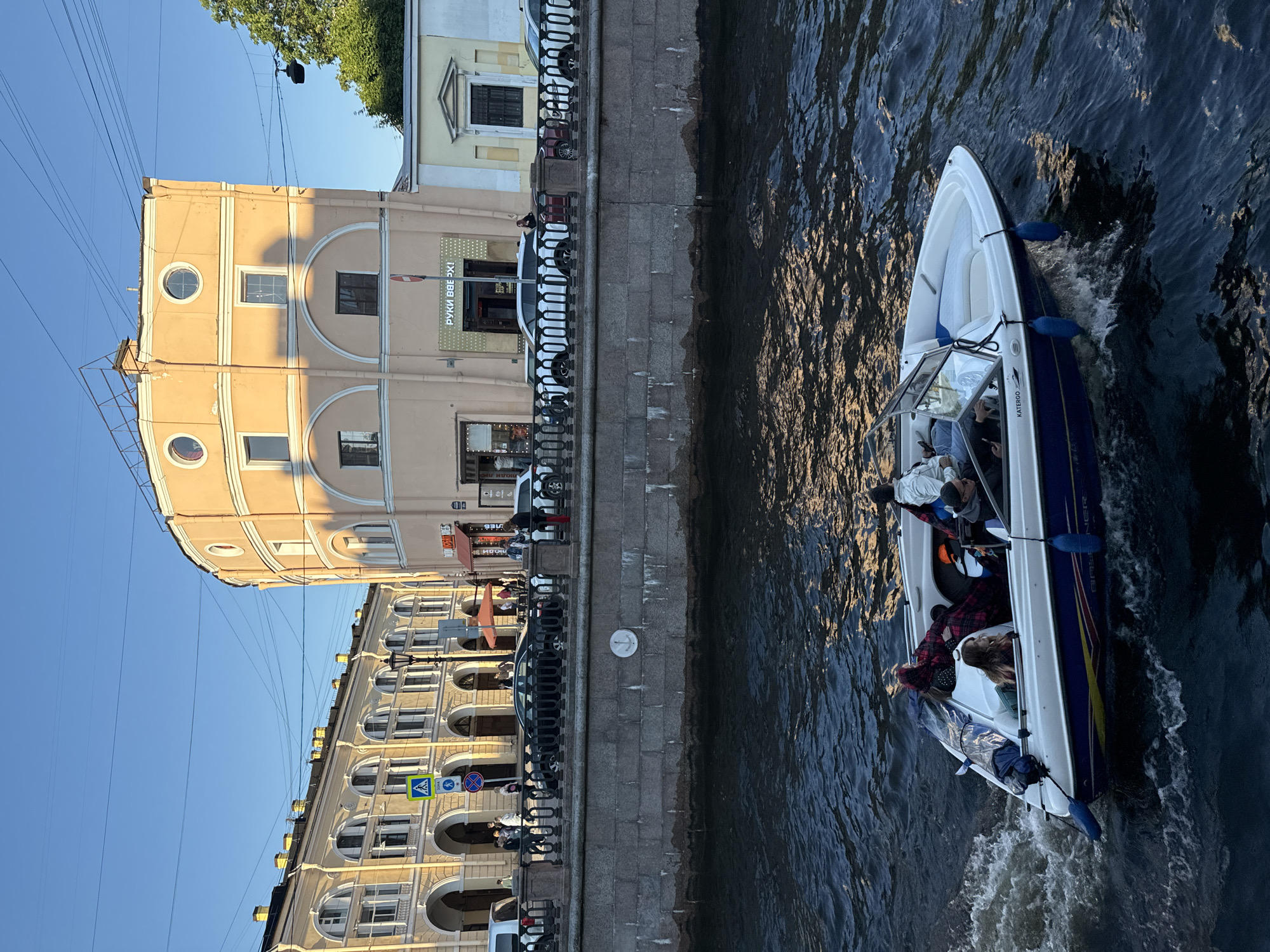 This is a low-angle, eye-level shot taken from a body of water, likely a canal or river, in Saint Petersburg, Russia. The foreground features a white and blue speedboat with several passengers navigating the choppy, dark water. The boat is moving from left to right, leaving a wake. The passengers appear to be casually dressed and engaged in conversation.
In the midground, a stone embankment separates the water from a city street lined with buildings. The buildings are characteristic of Saint Petersburg architecture, featuring classical facades with arched windows and ornate detailing. One prominent building is a light peach-colored, circular structure with arched windows and decorative circular accents on its upper level. Another building to the left is a pale yellow, with a colonnade of arches along its ground floor. The street is busy with pedestrian traffic and several cars are parked or in motion. Traffic lights and street lamps are visible.
The background is dominated by a clear, bright blue sky. The overall lighting suggests it is late afternoon or early evening, with the sun casting long shadows and illuminating the facades of the buildings. The scene depicts a vibrant urban setting where leisure activities on the water intersect with the city's historic architecture.
