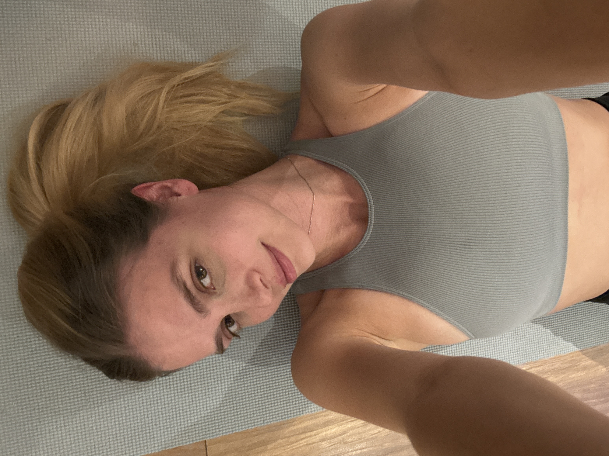 A woman with blonde hair pulled back in a ponytail lies on a gray mat, wearing a gray ribbed tank top. She is looking at the camera. The scene is indoors with wooden flooring visible in the lower right corner. The location in the city is Unknown, Unknown.