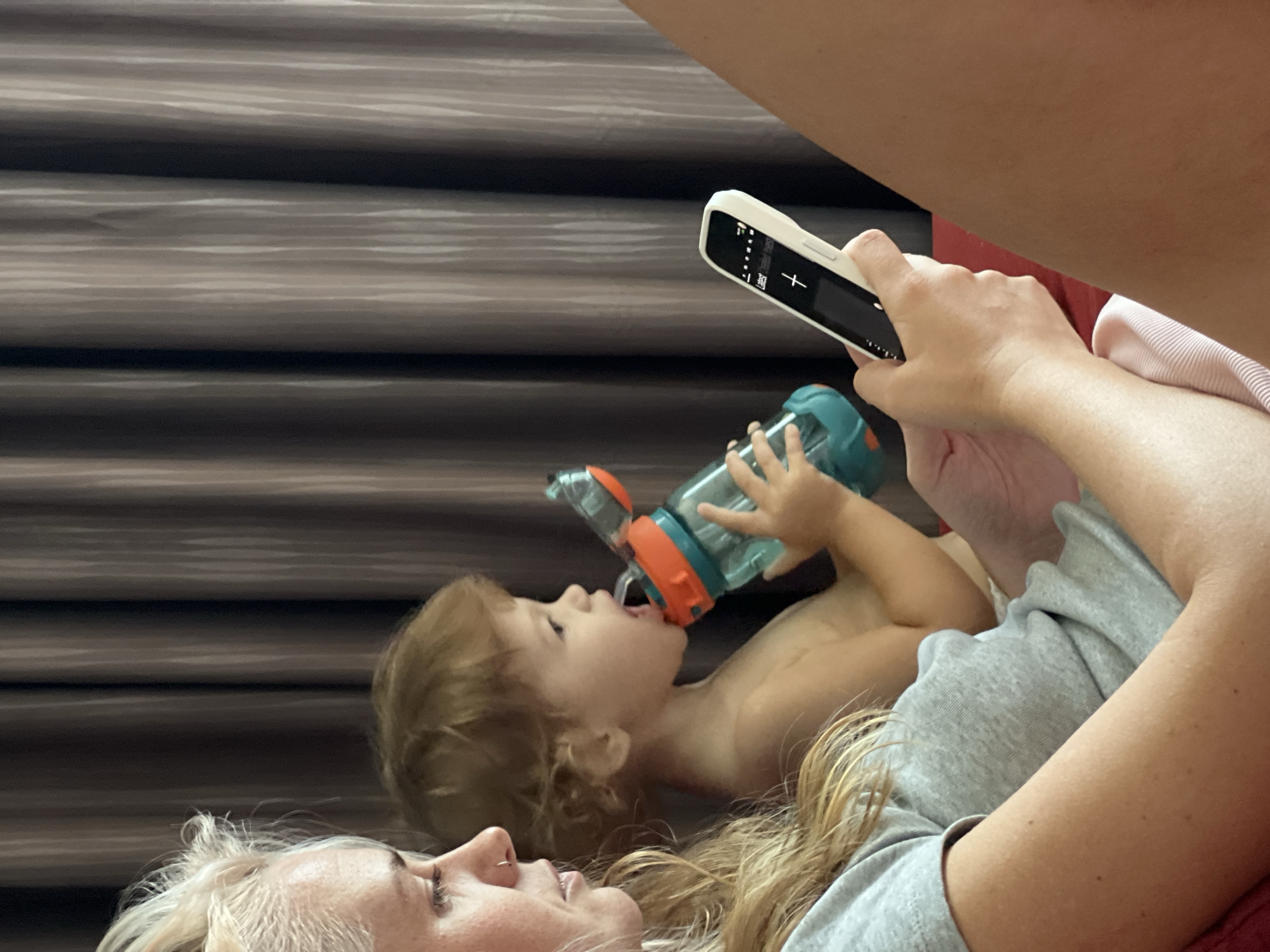 The image presents an intimate indoor scene featuring a woman, a child, and a third person's arm holding a smartphone. In the lower left foreground, an adult woman with blonde hair, seen from the chest up, lies reclined and looks towards the left with a calm expression. She has a small nose piercing and wears a grey t-shirt. Her long hair spreads across the surface she is lying on.
Adjacent to the woman, a young child, likely a toddler with light brown hair, is positioned above and to the right of her. The child is shirtless and lies on their back, actively drinking from a teal and orange sippy cup. Their small hands grip the cup firmly.
In the upper right section of the frame, a person's arm and shoulder are visible, holding a white-cased smartphone. The phone screen displays a dark interface with white elements, including the word "Lags," a "+" symbol, and several small icons, suggesting an application or photo editor is open. A pink and white striped fabric is visible beneath the phone.
The background consists of prominent horizontal dark and light brown stripes, resembling blinds or a textured wall, indicating an indoor setting. The overall mood of the scene is relaxed and domestic. No specific time of day or location cues are explicitly visible beyond the indoor environment.