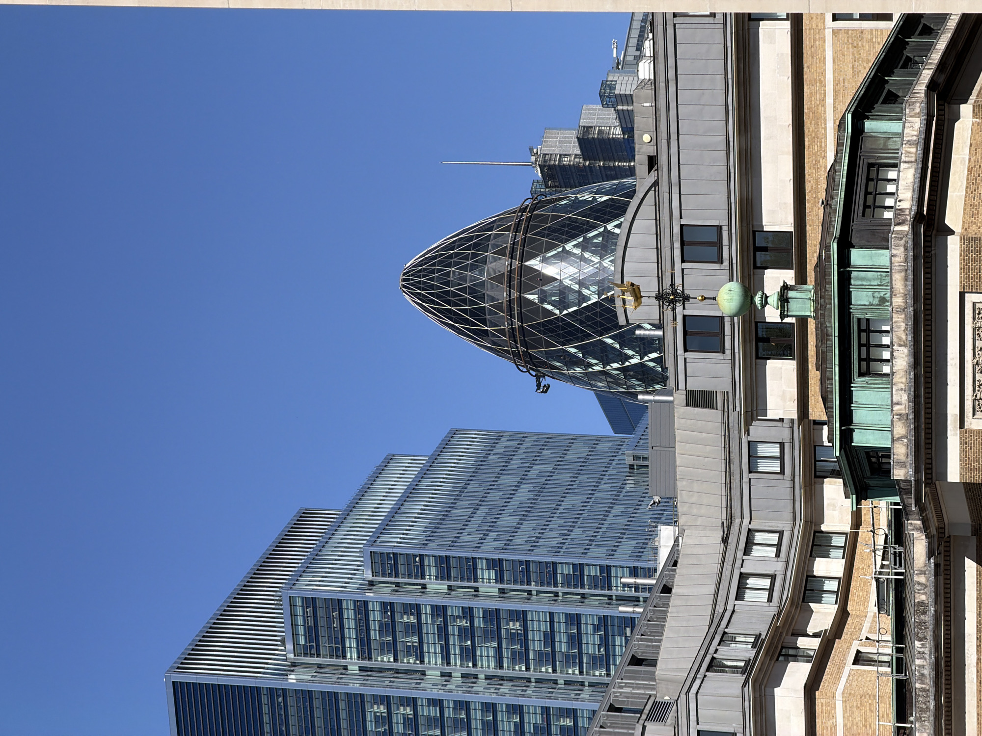 The image displays a clear, sunlit urban landscape in London, United Kingdom, featuring a blend of modern skyscrapers and traditional buildings. The most prominent subject is the iconic 30 St Mary Axe, known as The Gherkin, standing tall on the right side of the frame. Its distinctive, glass-paneled, diagrid structure reflects the bright blue sky.
To the left of The Gherkin, several other contemporary high-rise buildings are visible, characterized by their sleek, reflective glass facades and tiered architectural designs. These modern structures contrast with older, more traditional buildings in the foreground and lower right, which exhibit brickwork, stone elements, and details like a patinated green roof section. A decorative element resembling a golden grasshopper or weather vane is perched on an ornate dark structure on one of these older buildings, alongside a large spherical green finial.
The setting is a bustling financial district, with no specific activity or event taking place, presenting a static architectural scene. The time of day is clearly midday or early afternoon, indicated by the strong sunlight and distinct shadows. The weather is bright and clear, with a vivid, cloudless blue sky filling the upper portions of the image. No people or legible text are visible. The image captures the dynamic architectural diversity common in central London.