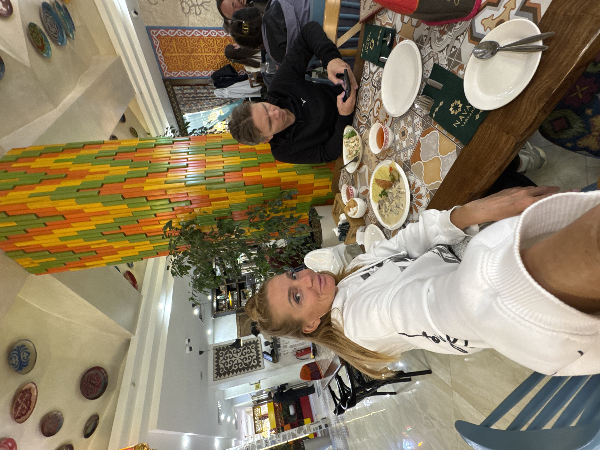 The image captures an indoor scene inside a restaurant or cafe, likely a "Chaikhana" as indicated by the visible text, with a distinctive Central Asian aesthetic. The overall setting is brightly lit, suggesting either daytime or well-illuminated evening.
In the foreground, a woman with blonde hair, wearing a white sweatshirt, looks directly at the camera with a slight smile. She is seated at a wooden table with a colorful, patterned surface, possibly tiled or a printed design. Across from her, a man with light brown hair, dressed in a black long-sleeved shirt, is engrossed in his black smartphone. Several white plates, bowls, cups, and cutlery are arranged on the table, along with various dishes including what appears to be a creamy meat and potato dish, a fresh salad, and small side dishes. Two dark green bags bearing the text "NAVA ЧАЙХАНА" (Nava Chaikhana) are also visible on the table.
The interior decor is rich and vibrant. One wall features a striking arrangement of horizontal rectangular tiles or blocks in alternating shades of orange, yellow, and green. Another wall is adorned with decorative, intricately patterned plates. Potted plants or small trees are strategically placed within the restaurant, adding greenery to the space. The ceiling is high, with visible light fixtures, and other patterned rugs or textile art pieces are hung on walls in the background, further enhancing the ethnic atmosphere. The general ambiance appears relaxed and pleasant, with the woman's smile contributing to this mood. Given the provided context, this establishment is located in Bishkek City, Kyrgyzstan.
