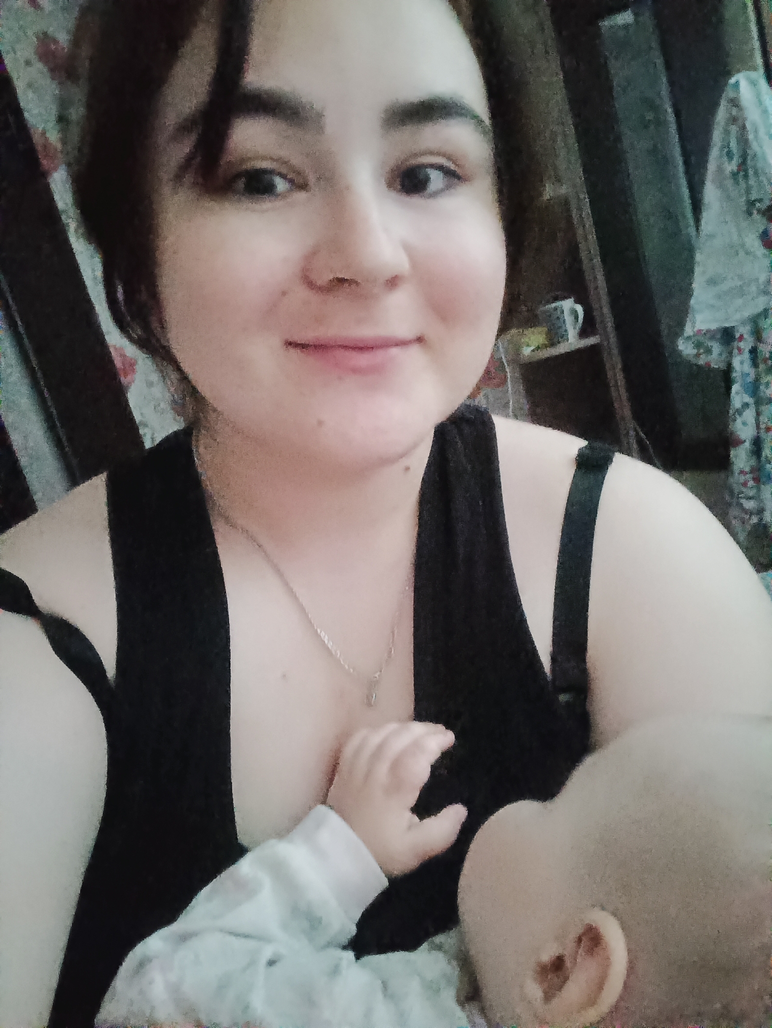 The image shows a woman with brown hair and a black tank top, smiling and looking directly at the camera. She is wearing a silver necklace. A baby, dressed in a light-colored, patterned onesie, is positioned close to her chest. The baby's head and one hand are visible. Behind the woman is a cabinet or shelf with a mug visible on it. Part of a patterned piece of clothing is also visible behind her. The location is not identifiable as Noviy, Russia from the provided image.