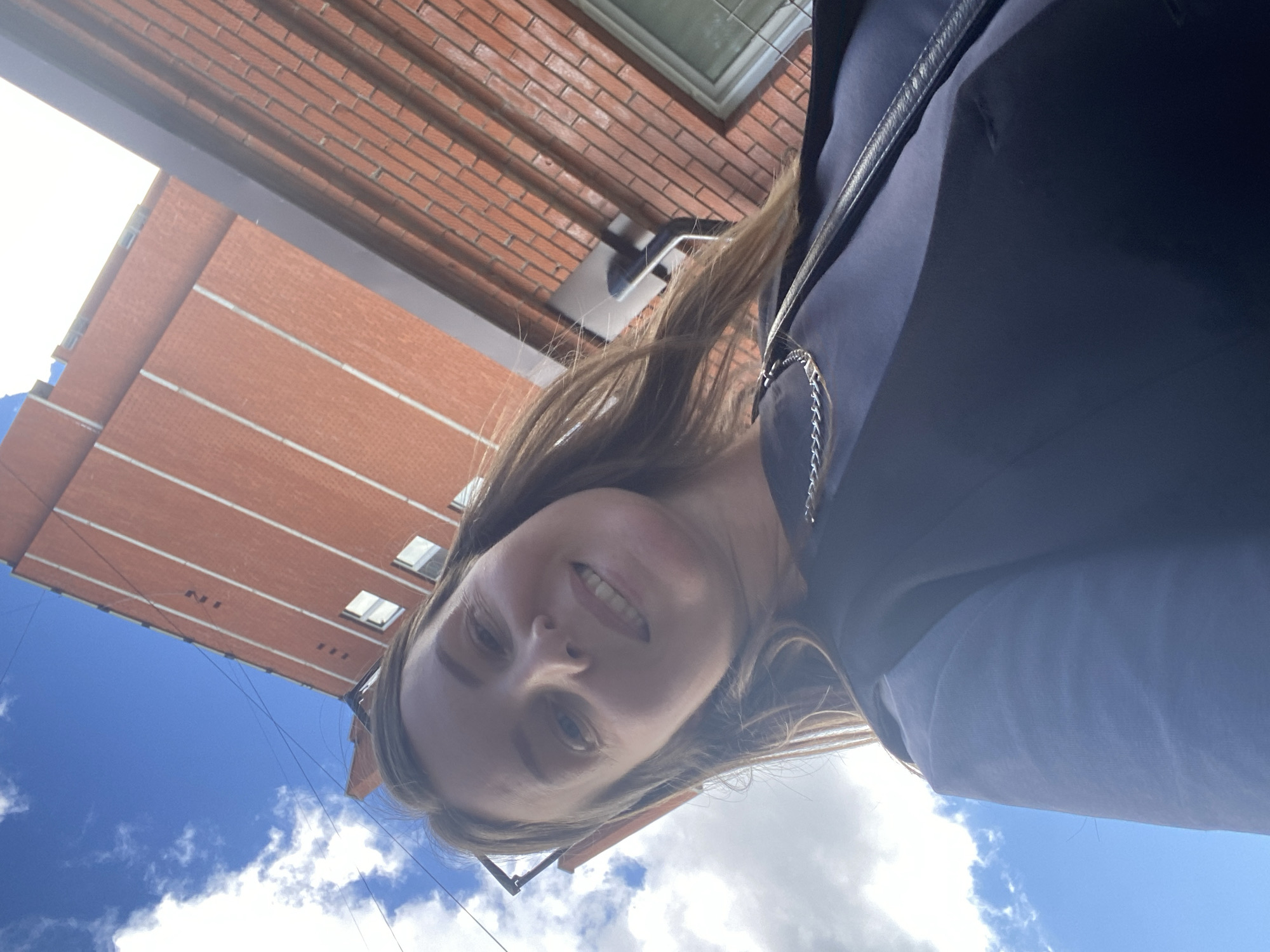 The image is a selfie taken outdoors in Chaikovsky, Russia, under a bright blue sky with scattered white clouds. The perspective is from below, looking up at the subject and a tall, brick building.
The subject is a woman with shoulder-length, light brown hair. She is smiling and looking towards the camera. She is wearing a dark top and a light blue shirt underneath. A thin silver chain is visible around her neck. Her sunglasses are perched on her head.
The building behind her is made of red brick with horizontal white bands. Several windows are visible on the building. A dark metal gutter or pipe runs down the side of the building. The overall impression is that of an urban setting, possibly residential or commercial.