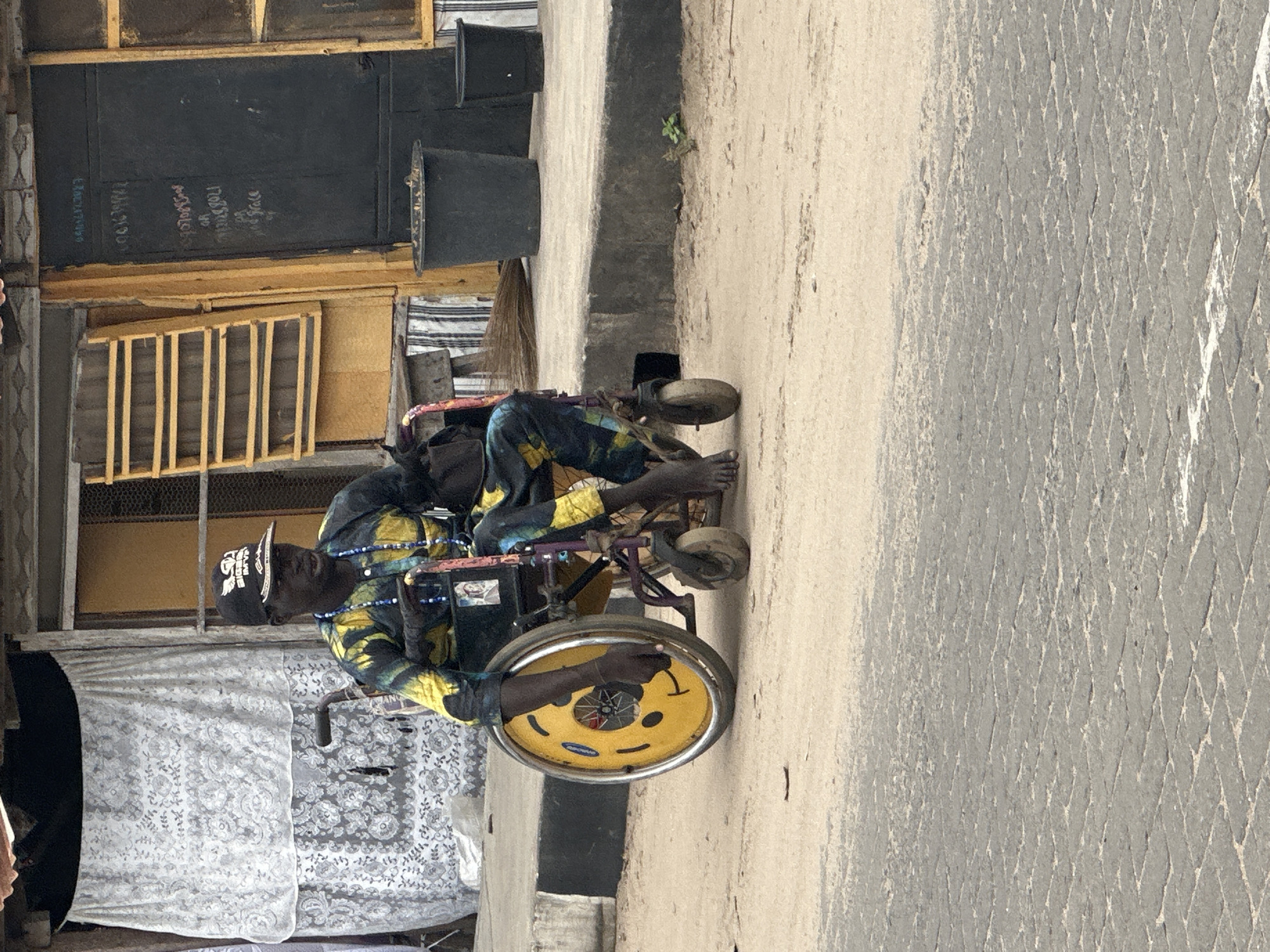 In Badagry, Nigeria, a person in a wheelchair with a smiley face sticker on the wheel is seen from the side. The individual is wearing a patterned shirt and pants, a cap, and beaded necklaces. The wheelchair is a standard manual model with a yellow wheel decorated with a smiley face. The background shows a building with a yellow door and window, a white curtain hanging outside, and some potted plants. The ground is paved with bricks. The image captures a moment of everyday life in this Nigerian city.