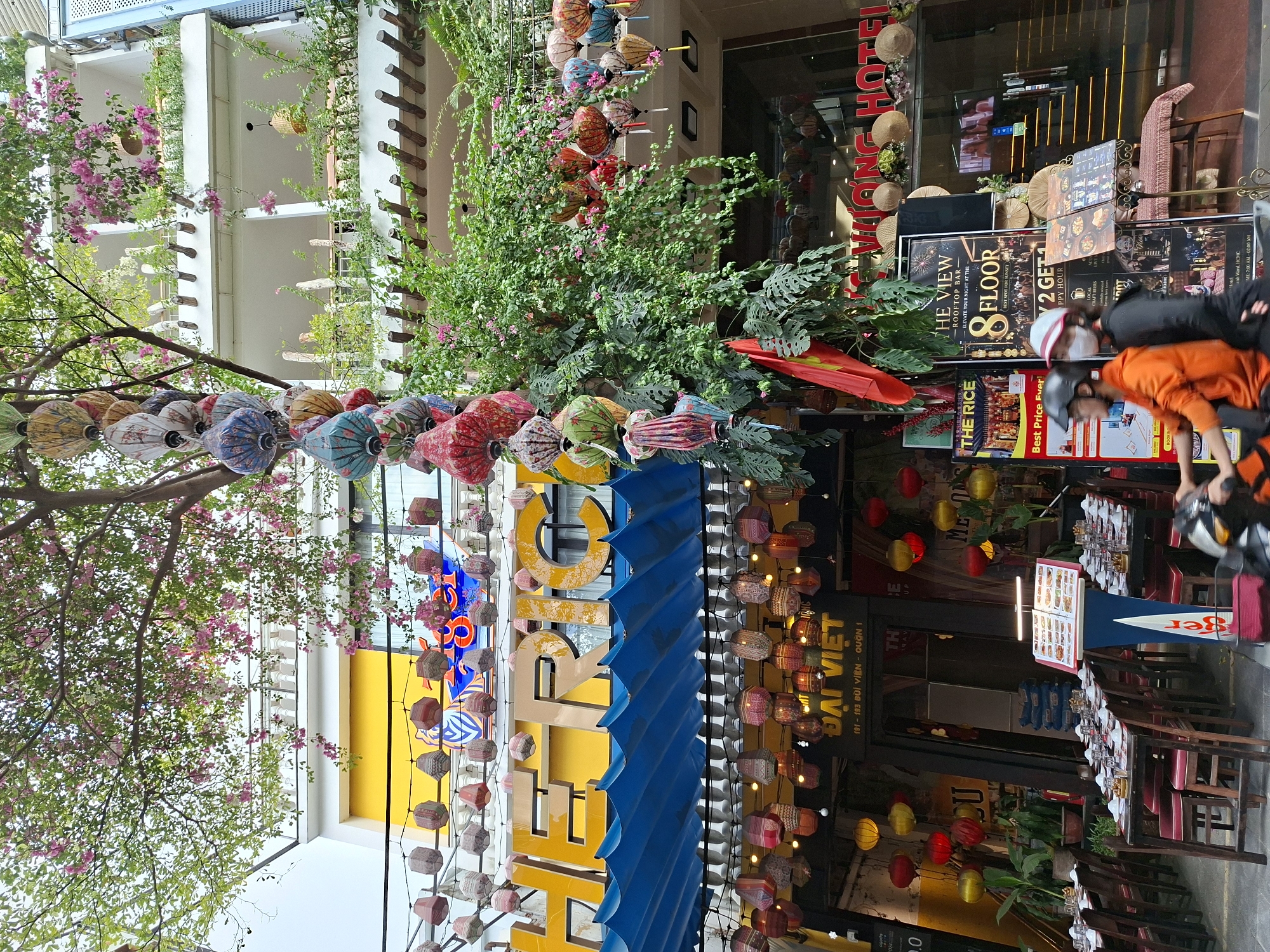 The image captures a street view in Thủ Đức, Vietnam, dominated by a building adorned with numerous colorful lanterns and lush greenery. The building features a yellow facade on the upper levels with white balconies, draped with flowering vines. Below, a blue awning covers a row of outdoor seating at a restaurant. The street-level features more decorative lanterns, a Vietnamese flag, and signage including "THE RICE" and "THE VIEW ROOFTOP BAR". Two individuals on a scooter are passing by. Several tables with white tablecloths and place settings are visible in front of the restaurant, indicating it is open for business. The overall scene suggests a vibrant and commercial street in Vietnam.
