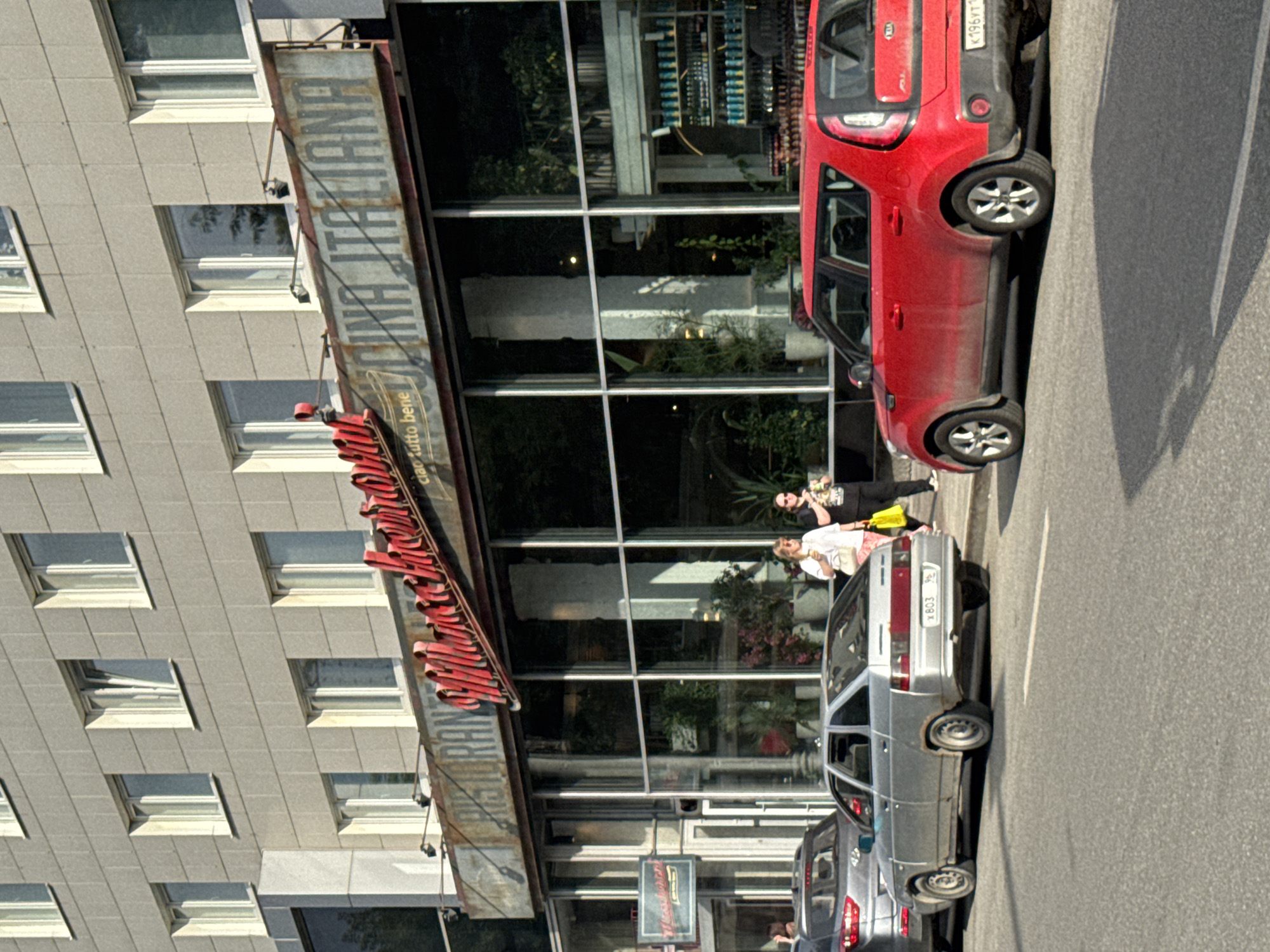 The image shows a street scene in Yekaterinburg, Russia, likely in an urban area with a multi-story building. In the foreground, a red Kia Soul is parked on the right side of the road, and a silver sedan is parked to its left. Both vehicles are facing away from the viewer.
The building behind the cars features a prominent sign for "RISTORANTE Cucina Italiana" in a retro, slightly distressed style. The restaurant has large, reflective windows that show greenery and interior pillars. Two women are walking on the sidewalk in front of the restaurant. The woman on the left is wearing a white top and pink pants, carrying a white bag. The woman on the right is wearing a black top, dark pants, and sunglasses, carrying a yellow bag.
The street is paved and appears to be a typical city road. The building's facade is clad in grey stone or concrete panels. The overall impression is a sunny day in a commercial or mixed-use district.