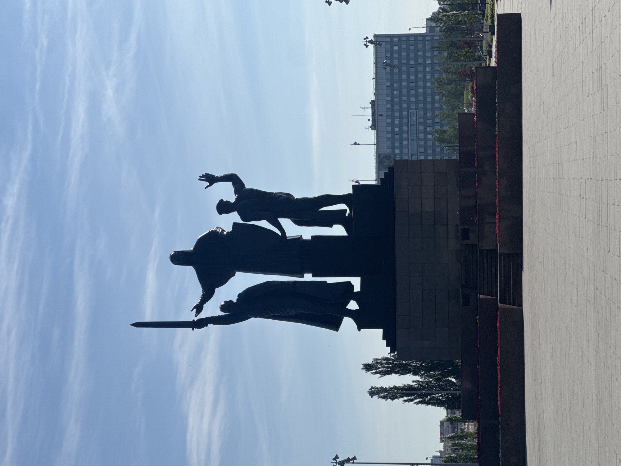 A large, dark-colored memorial monument dominates the left side of the frame, silhouetted against a bright blue sky. The monument features three heroic, stylized human figures in dynamic poses, standing on a multi-tiered, angular base. The figure on the viewer's left points a sword forward and upward, while the central figure reaches out with an arm, and the figure on the right raises an arm towards the sky. The sculpture evokes themes of struggle, heroism, and remembrance.
In the background, behind the monument, a modern, multi-story building with numerous windows rises against the sky, accompanied by scattered green trees. A small Russian flag is faintly visible on a flagpole near the building. To the right of the monument and extending into the foreground, a wide, light-colored paved plaza or walkway is visible, made of square tiles. Dark, rectangular planters or structures, containing small red flowers, separate parts of the paved area from the monument's base.
The photograph was taken on a bright, clear day, with a blue sky and wispy white clouds, casting the monument into a strong silhouette. No discernible text is visible. The scene suggests a public memorial space in Perm, Russia.