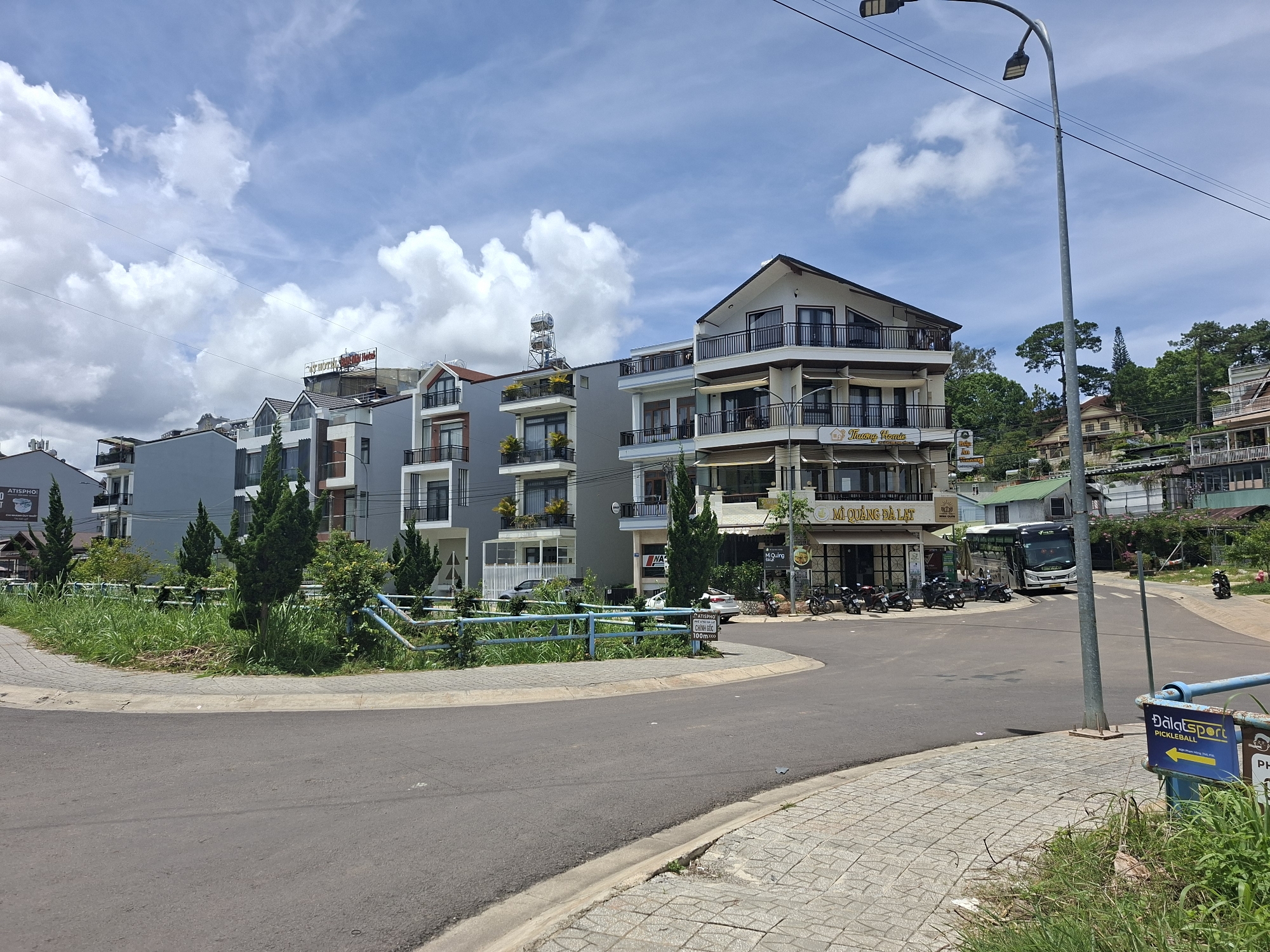The image depicts a street scene in Da Lat, Vietnam, featuring a cluster of modern multi-story buildings with balconies, characteristic of the city's architecture. The buildings are primarily light-colored, with some featuring grey facades and red-tiled roofs. Trees, including cypress and other evergreens, are planted along the roadside and in front of the buildings. A blue metal fence separates a grassy area from the paved road. A street light with two lamps stands prominently on the right side of the frame. Parked motorcycles and a bus are visible on the road. A sign with yellow lettering on a blue background, indicating "Dalatsport PICKLEBALL," is positioned near the foreground. The sky is partly cloudy, with white clouds against a blue backdrop. The overall scene suggests a bright, sunny day.