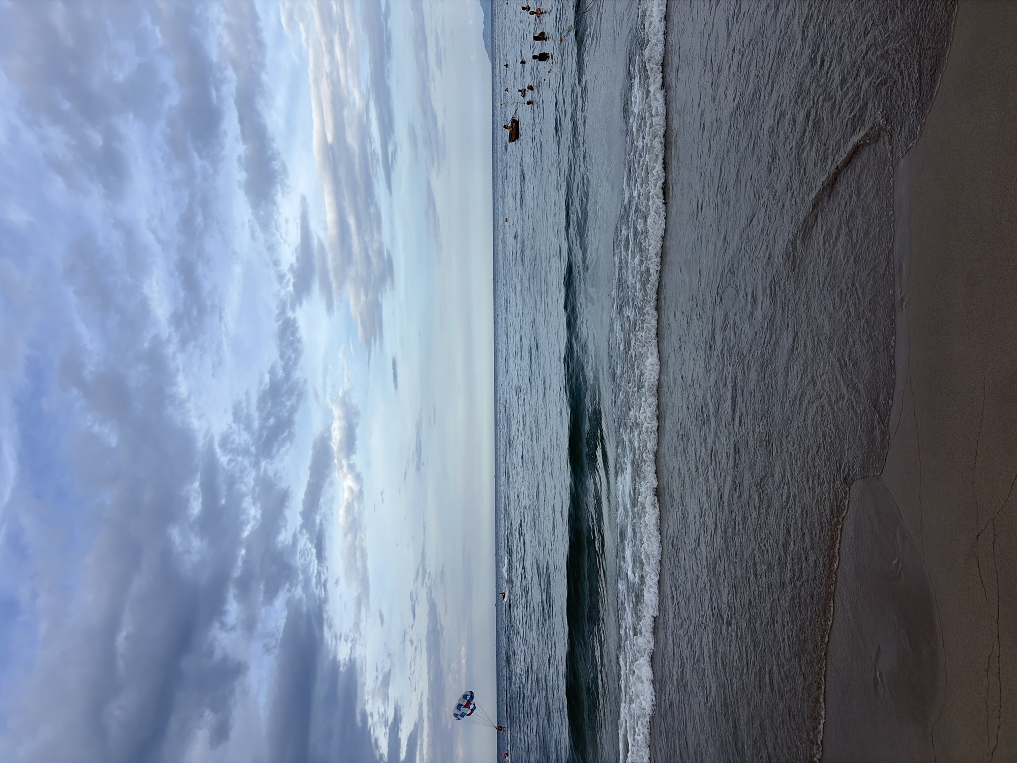 The image depicts a serene beach scene at My Khe Beach, Da Nang, Vietnam. The sky is overcast with dramatic, grey clouds interspersed with patches of lighter blue. Gentle waves are rolling onto the sandy shore, creating a foamy white crest. In the distance, several people are visible in the ocean. On the left side of the frame, a person is parasailing, a colorful parachute trailing behind them. Further out, a small orange boat is present with at least one person aboard. Other individuals are scattered throughout the water, some appearing to be swimming. The overall atmosphere is calm and slightly moody due to the cloudy sky.