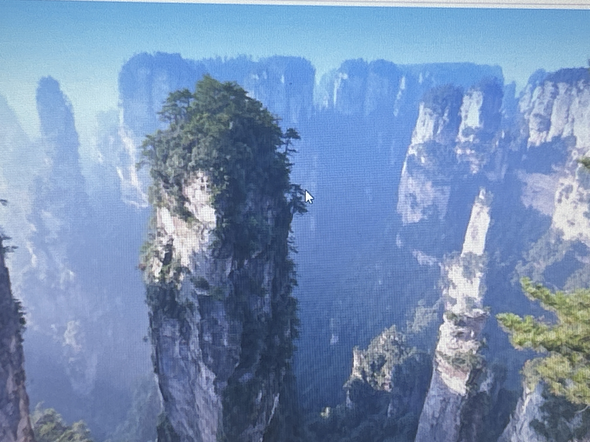 The image depicts a panoramic view of the Zhangjiajie National Forest Park in China, characterized by towering sandstone pillars covered in lush vegetation. The scene is bathed in a soft, ethereal light, with mist swirling between the colossal rock formations, creating a dreamlike atmosphere. The foreground features a prominent, elongated pillar, its rugged surface partially obscured by dense green foliage. In the background, a series of similar pillars recede into the hazy distance, their forms softened by the atmospheric conditions. The sky above is a clear, pale blue. There are no people, animals, or man-made structures visible in the image. The provided crops do not alter the content of the original image. The image is not related to Sofia, Bulgaria.