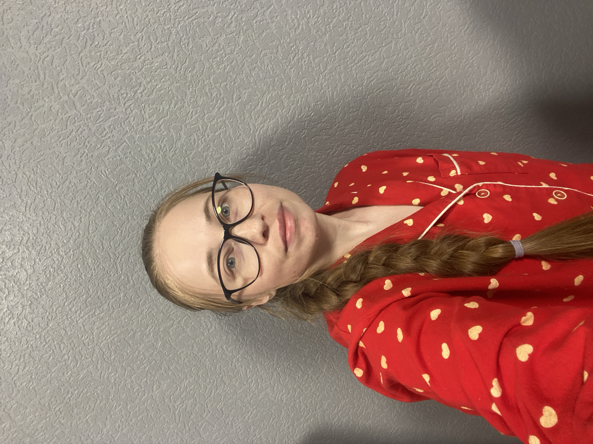 A woman with blonde hair braided and wearing glasses and a red and white heart-patterned pajama top is positioned in front of a textured gray wall. The image appears to be taken indoors, possibly a private residence. There is no indication of the location being Saint Petersburg, Russia or any other city.