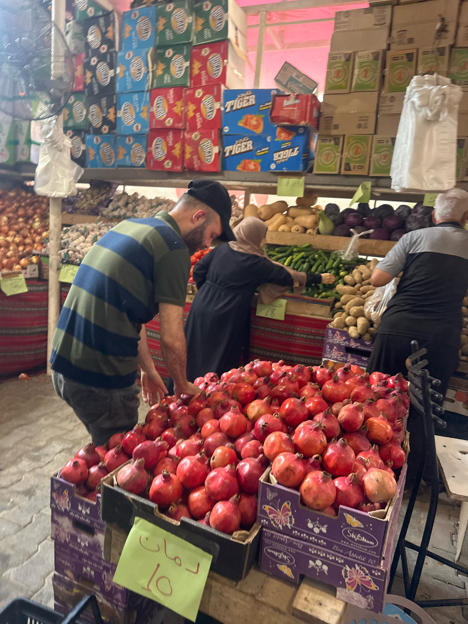 In the city of Gaza, Palestinian Territories, a market scene unfolds with a focus on produce. In the foreground, two stacked cardboard boxes overflow with pomegranates. A yellow sign with Arabic writing and the number "10" is attached to the front of the top box. Behind the pomegranates, a man in a green and blue striped shirt and dark pants is visible, his back to the camera, reaching towards the fruit. To his right, a woman wearing a hijab and black abaya is selecting cucumbers from a crate. Beside her, another man in a grey and black shirt is facing away, looking towards the produce. In the background, shelves are stacked with boxes of various goods, including some with the "Tiger" logo. Further back, piles of onions and other produce are visible, along with a large fan. The overall impression is a bustling market atmosphere.