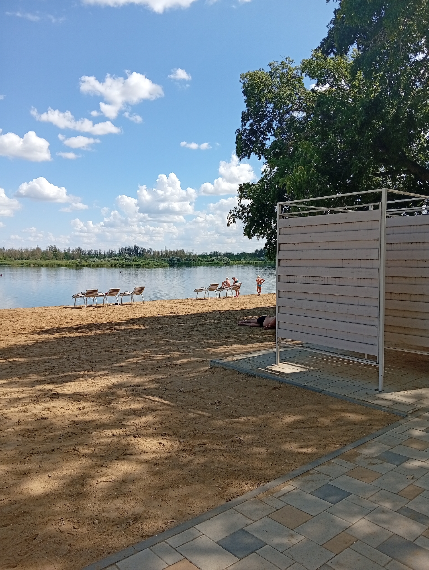 The image depicts a sandy beach area next to a body of water, likely a lake or river, under a bright blue sky with scattered white clouds. In the foreground, a changing room structure with horizontal wooden slats stands on a paved area that transitions to sand. To the left of the changing room, several people are visible on the beach. One person is lying on the sand near the changing room. Further back, closer to the water's edge, a group of people are sitting on lounge chairs, and another person is standing near the water. The water is calm and reflects the sky and surrounding trees. The shoreline is lined with trees and vegetation in the distance. The overall scene suggests a recreational area on a sunny day. The location is identified as Мичурин ауылдық округі, Kazakhstan.