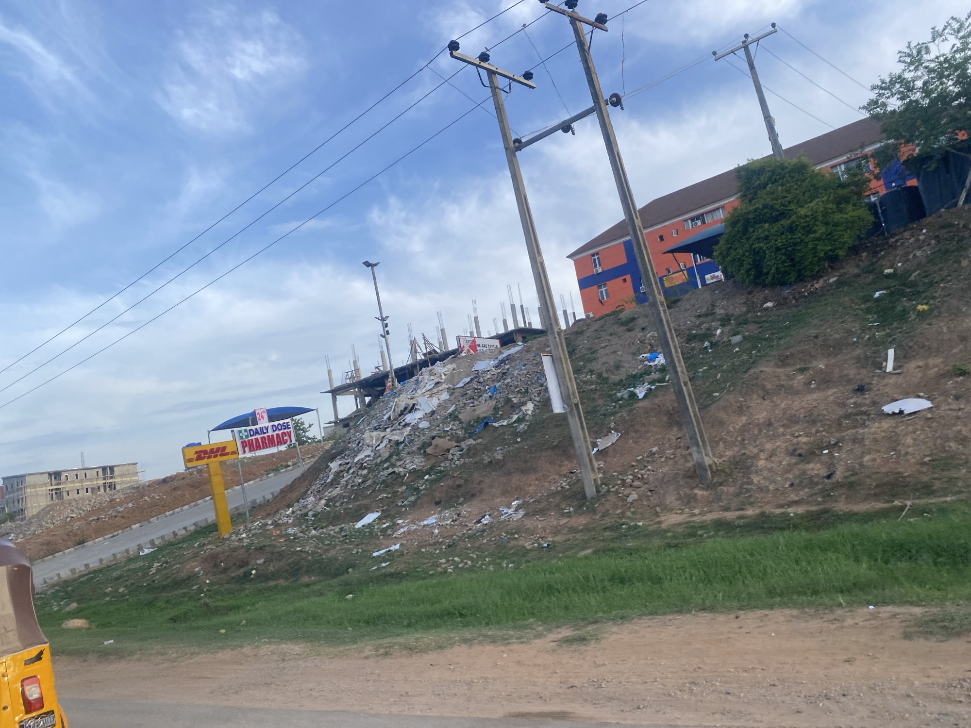 The image depicts a street scene in Abuja, Nigeria, with a view of a DHL Daily Dose Pharmacy sign and associated structures, situated on a dirt slope. In the background, there are unfinished buildings and a building with an orange and blue facade, and utility poles with wires. The slope is covered in trash and debris. The sky is partly cloudy. The foreground includes a partially visible yellow tricycle vehicle.