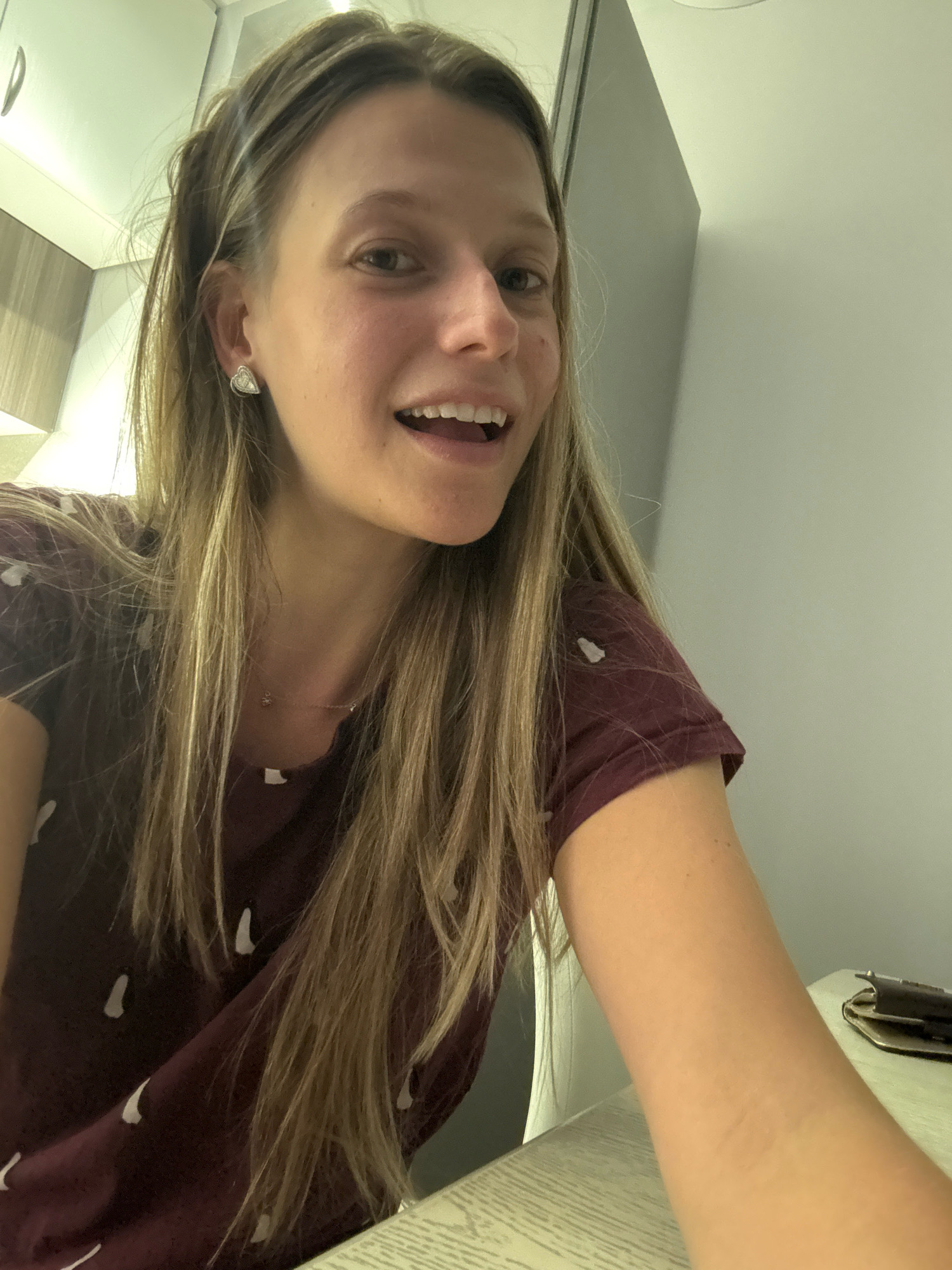 A young woman with blonde hair is taking a selfie in a well-lit room. She is wearing a dark red t-shirt with a subtle pattern. She has a heart-shaped earring in her left ear and a delicate necklace. Her mouth is open, revealing her teeth, and she appears to be smiling or laughing. The background is a light-colored wall and a glimpse of cabinetry. The scene suggests an indoor setting, possibly a home or a hotel room, and there is no indication of the city of Sofia, Bulgaria, or any specific urban environment.