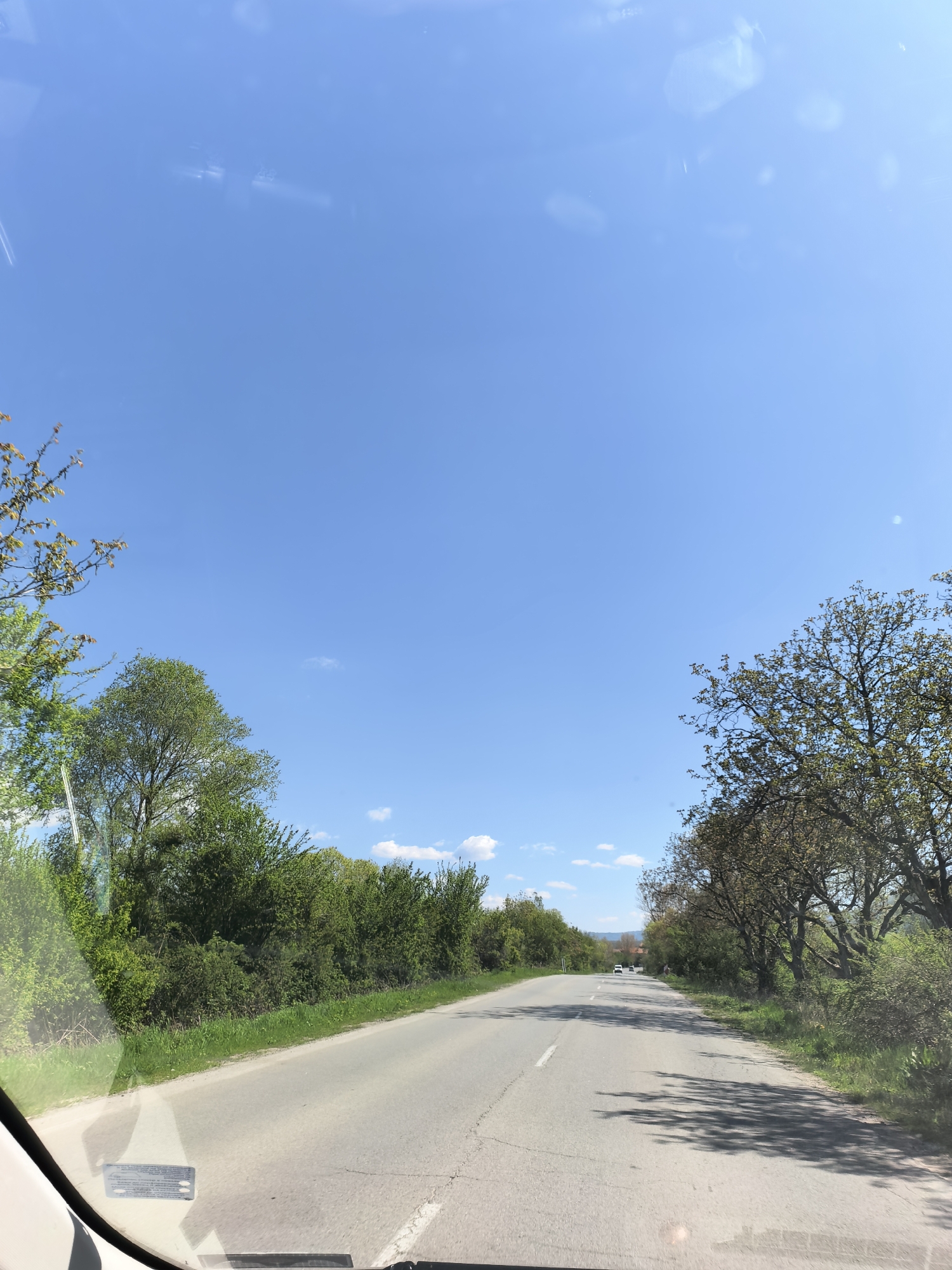 The image captures a first-person view from inside a vehicle, looking out through the windshield onto a rural road on a bright, sunny day. The road, made of asphalt with some cracks, stretches ahead and gently curves to the right, with a double white line marking the center and a solid white line on the right edge.
Lush green vegetation, including tall trees and dense bushes, lines both sides of the road. The trees on the right cast long shadows across the road, suggesting the sun is high in the sky, likely mid-morning to early afternoon. In the distance, several cars are visible on the road, indicating traffic. Beyond the greenery and distant road, hills or mountains can be seen under the clear blue sky, which has a few scattered white clouds.
The top portion of the image shows a vast expanse of clear blue sky. Reflections and some glare are visible on the windshield, especially in the upper section. The bottom part of the frame reveals the dashboard or interior of the vehicle, and a sticker or label is affixed to the lower left side of the windshield, containing unreadable text. The overall scene depicts a peaceful drive through a green, open landscape under pleasant weather conditions.